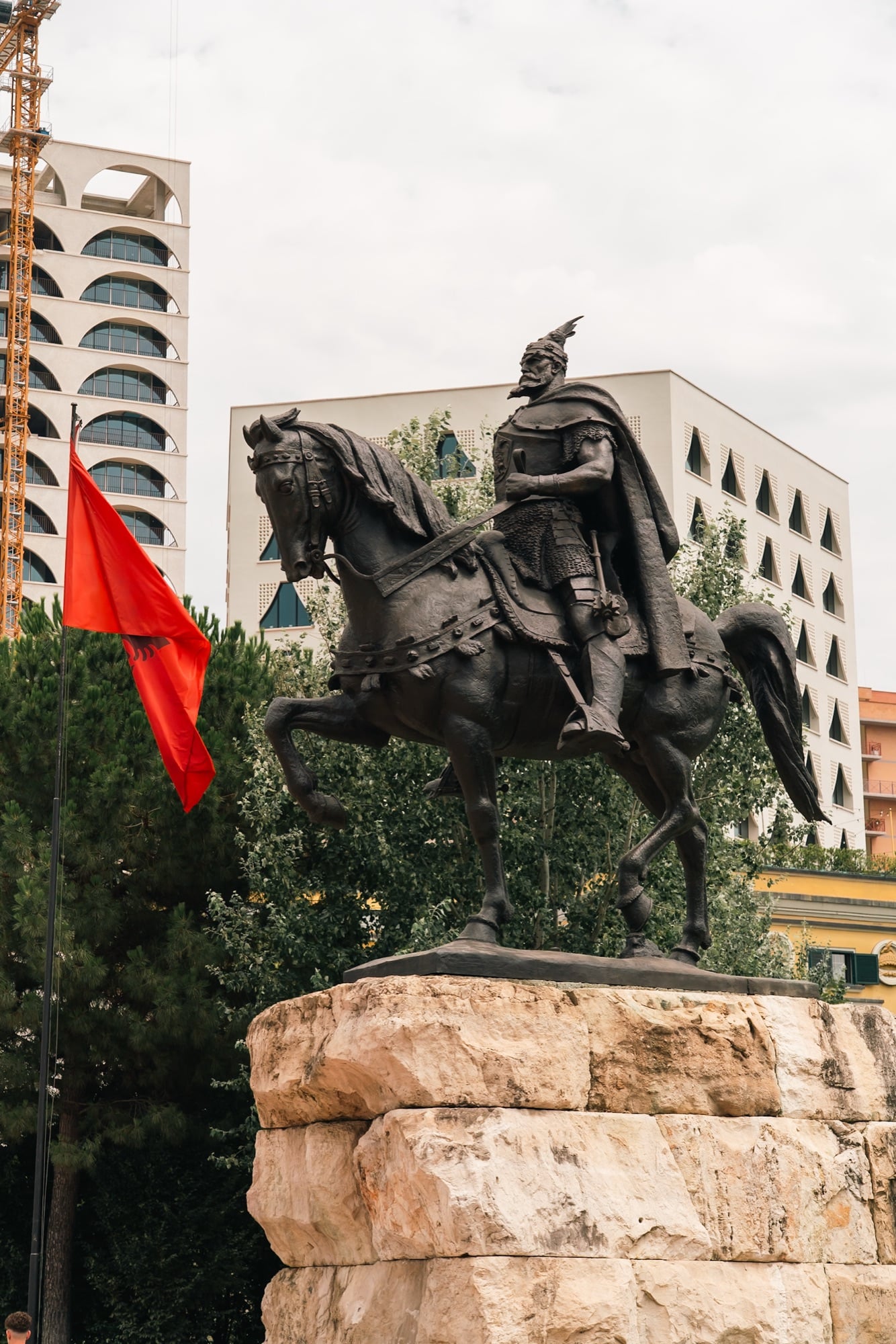 The bronze equestrian statue of Skanderbeg beside a red Albanian flag, city buildings rising behind