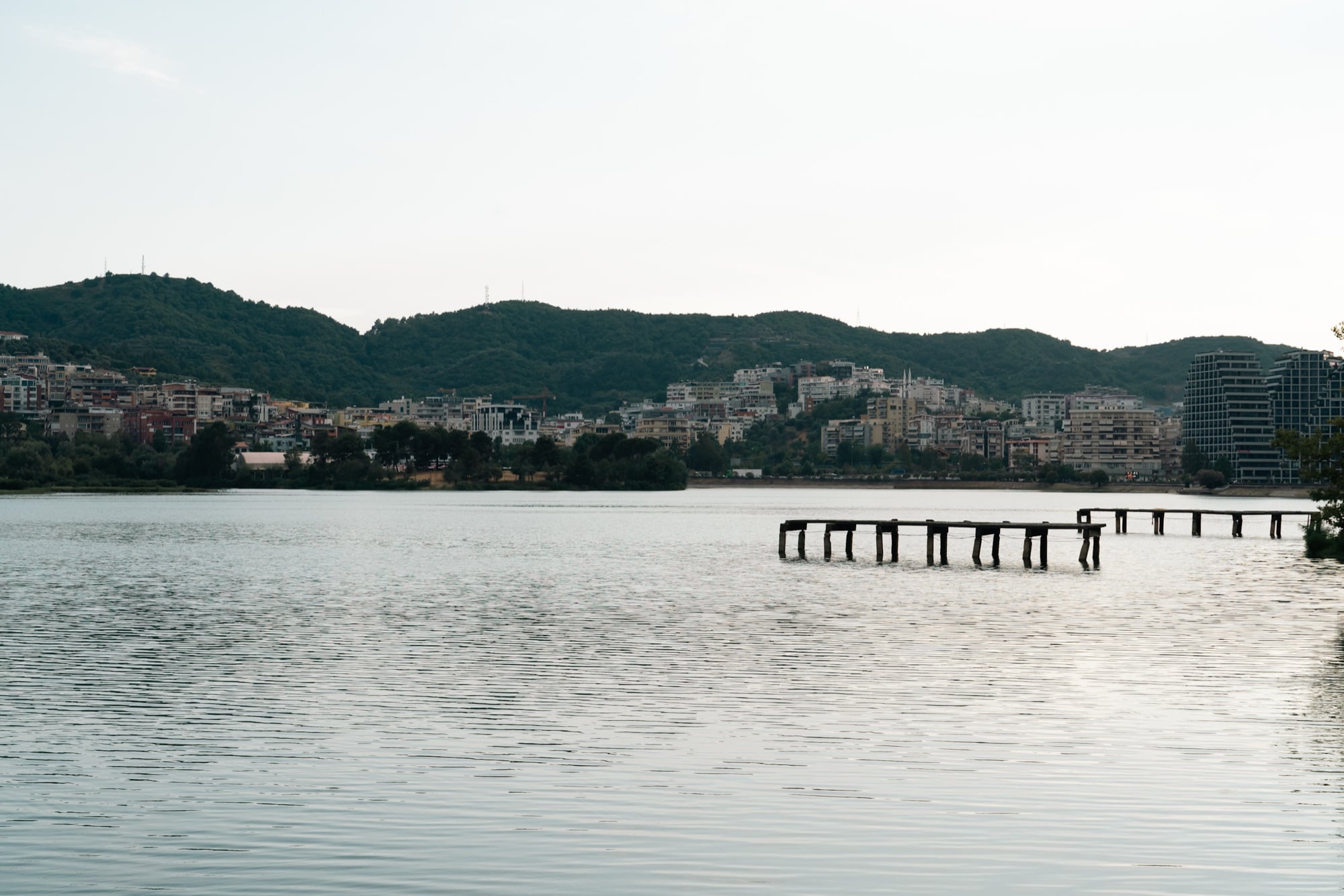 A weathered wooden pier reaching across the calm Artificial Lake of Tirana toward the city's hillside buildings