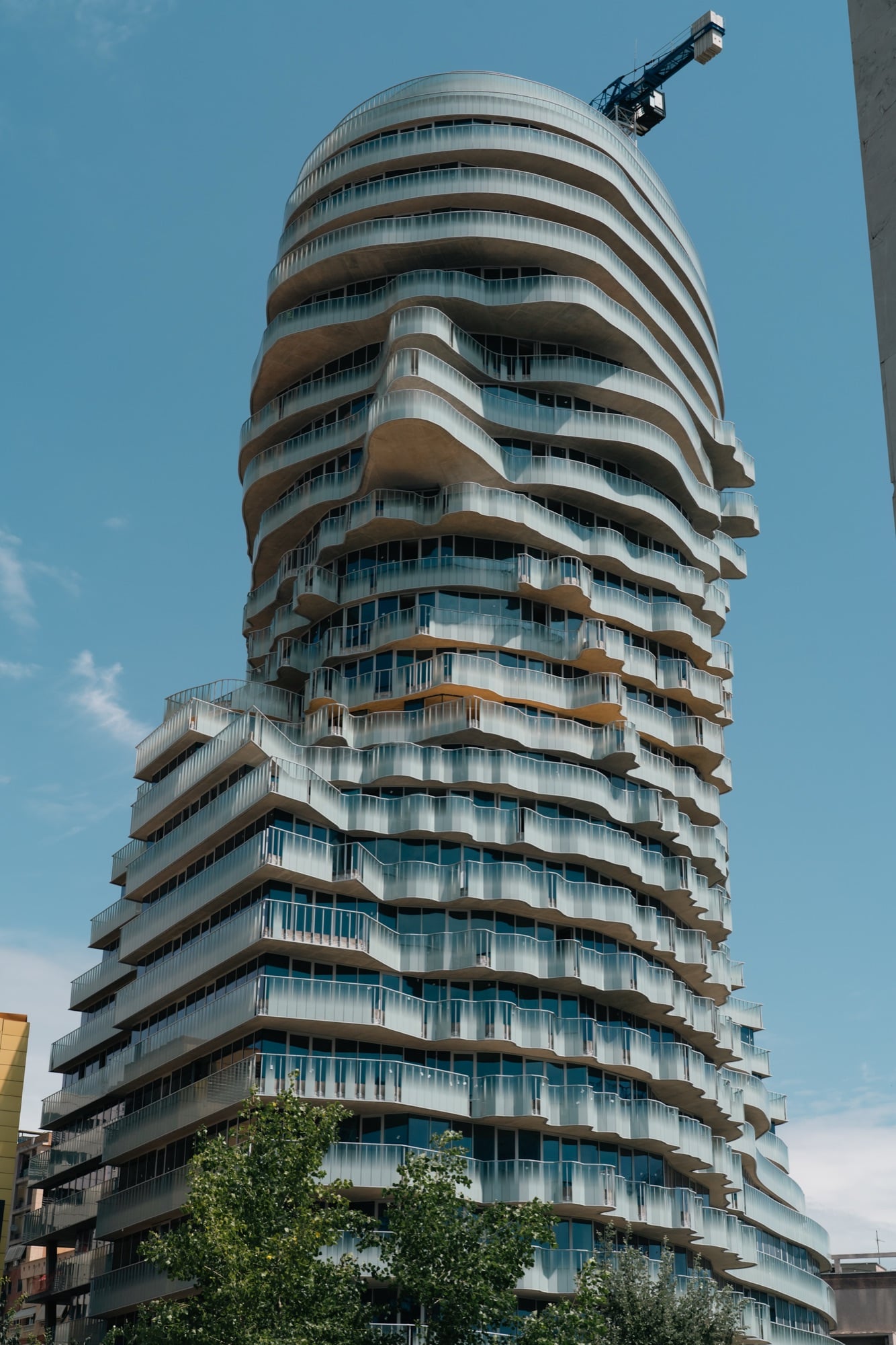 A residential tower of stacked wave-edged glass balconies rising against the blue sky, a crane at its crown