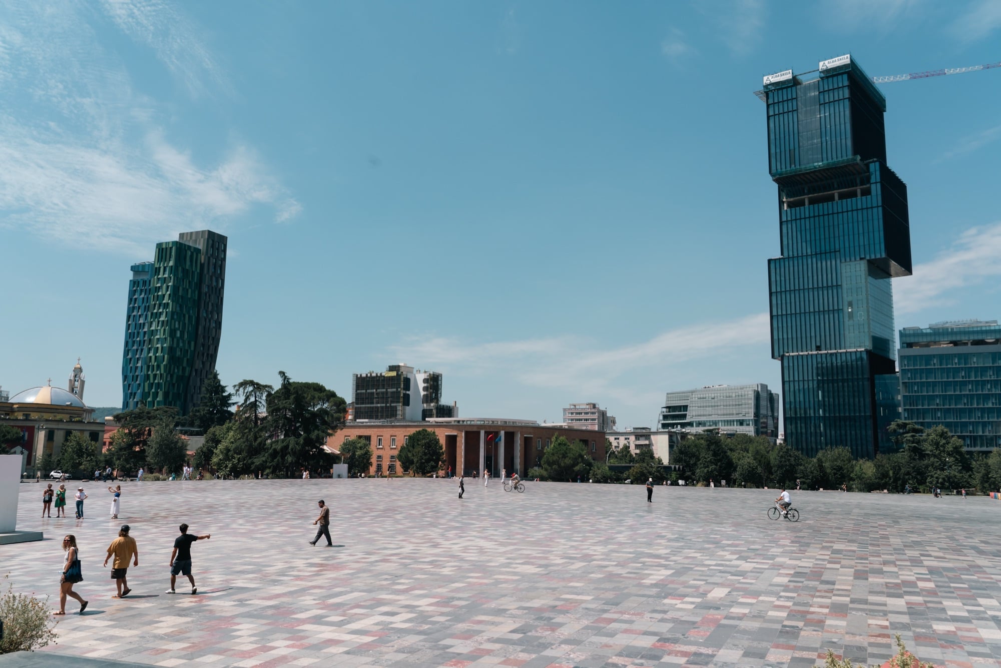 A wide view across the chequered paving of Skanderbeg Square, glass towers on the skyline and people crossing on foot and by bike