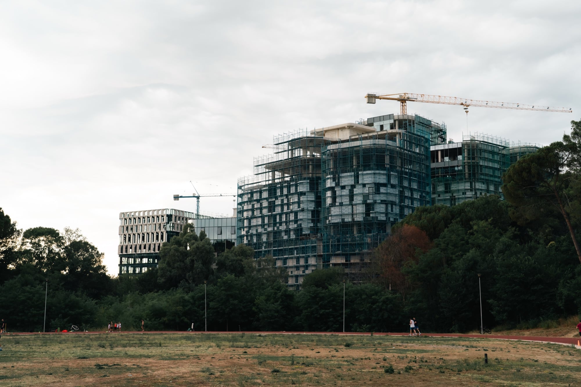 Scaffolded high-rise towers under construction rising behind a red running track and dry field, cranes overhead