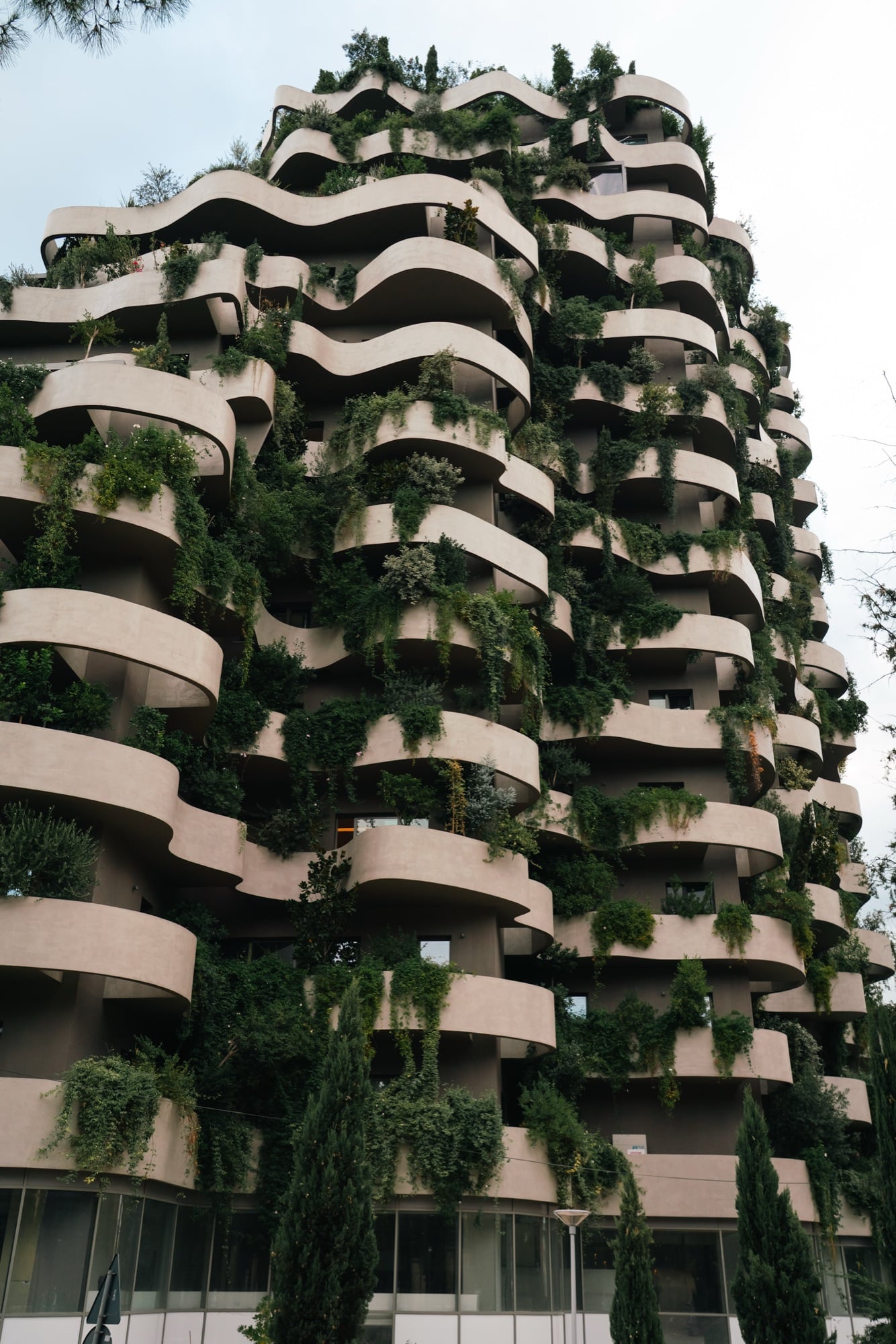 A residential tower with undulating balconies overflowing with greenery and cypresses, a vertical garden facade