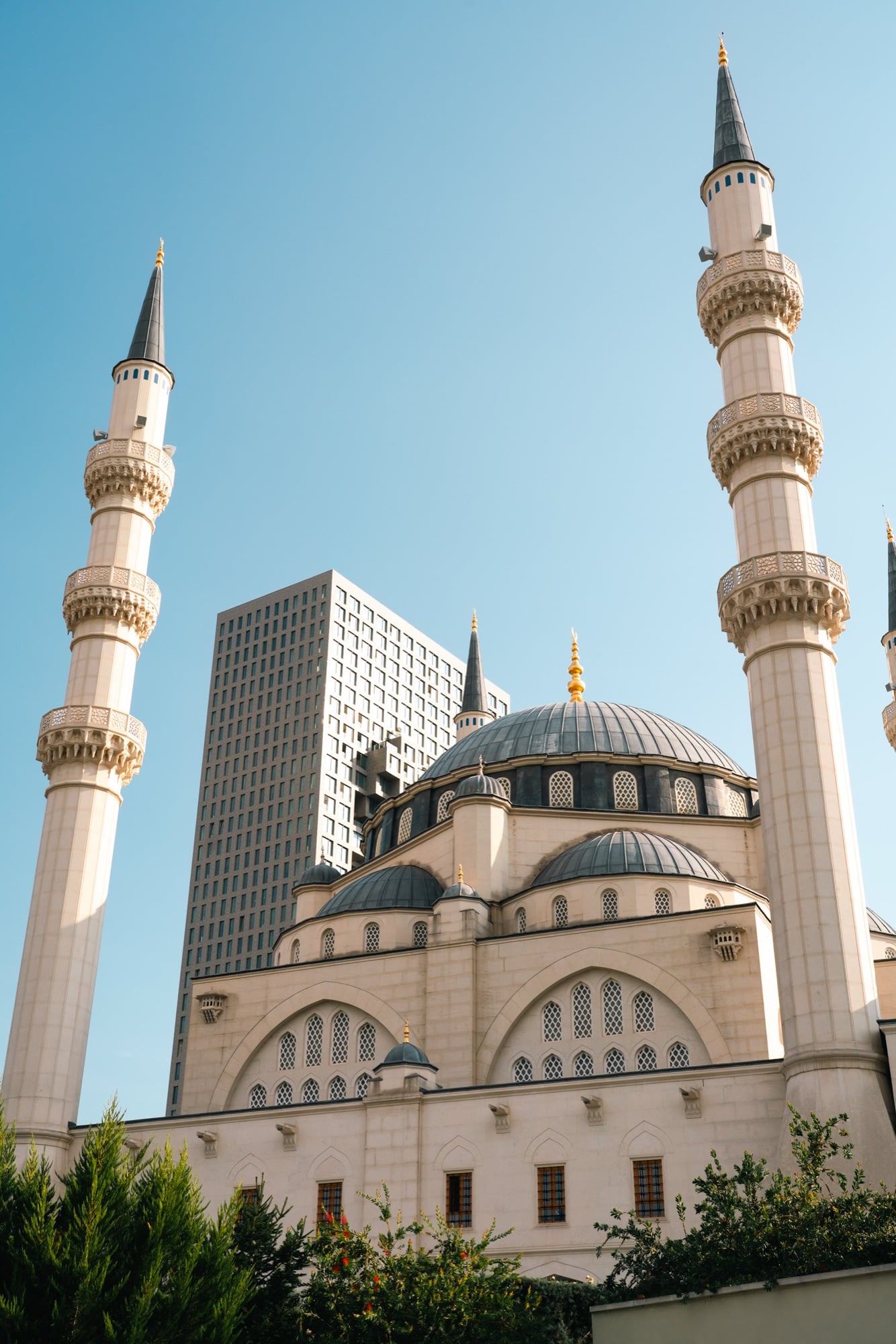 The twin minarets and lead-grey domes of the Great Mosque of Tirana rising against a clear sky, a modern tower behind
