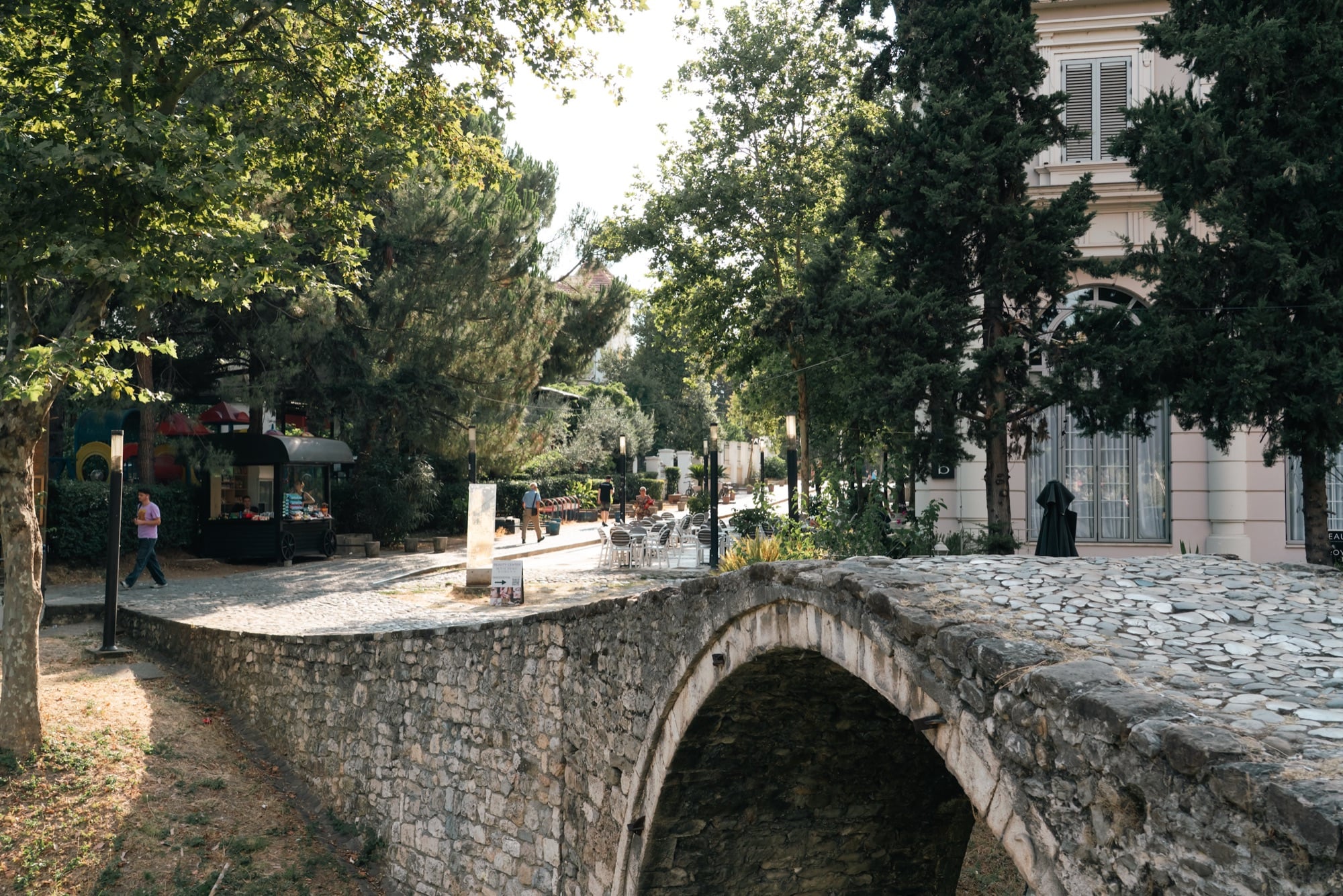 The old Ottoman Tabak stone bridge arching over a dry bed in a leafy park, a café kiosk and passers-by beyond