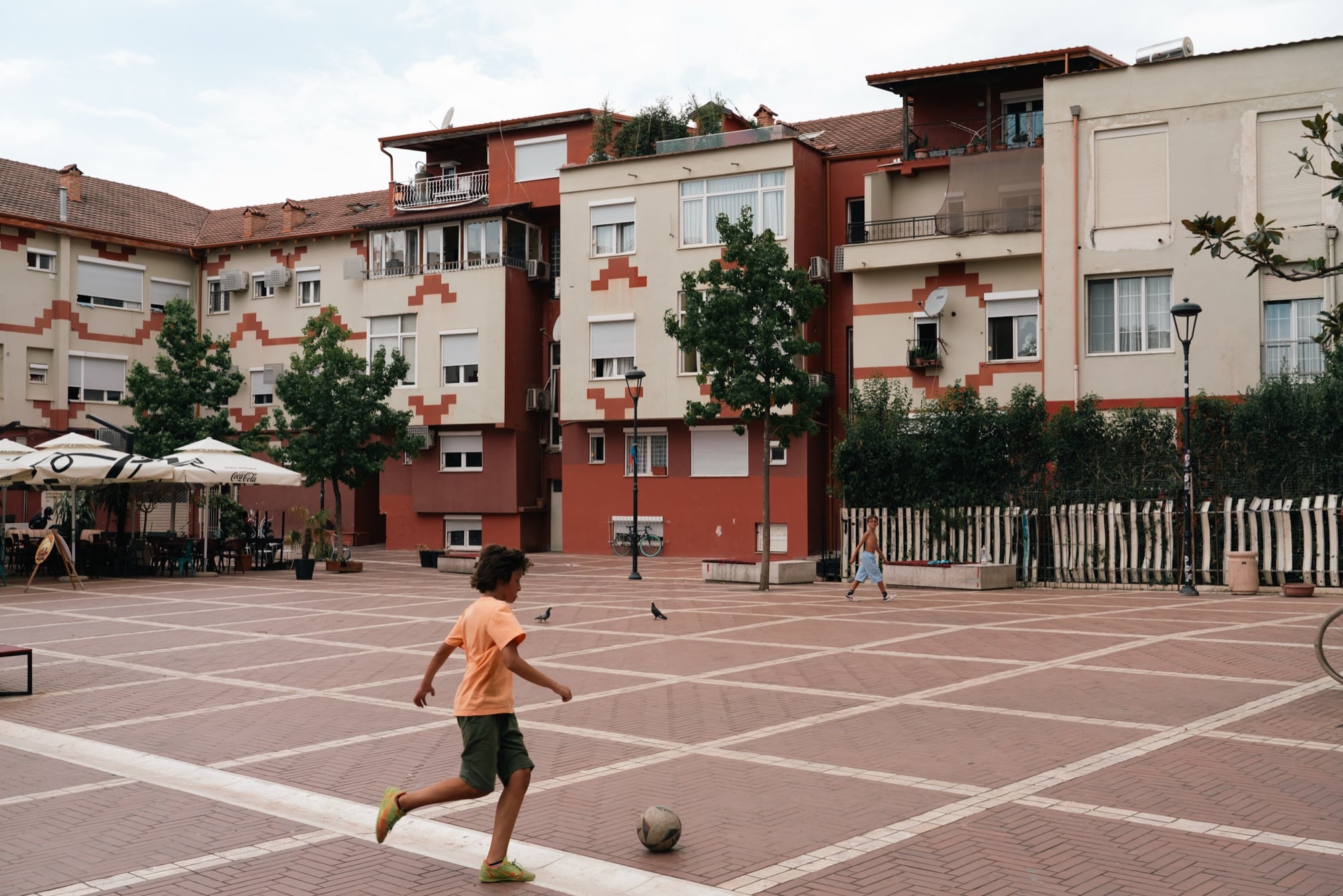 A boy dribbling a football across a red-tiled courtyard ringed by cream-and-terracotta apartment blocks