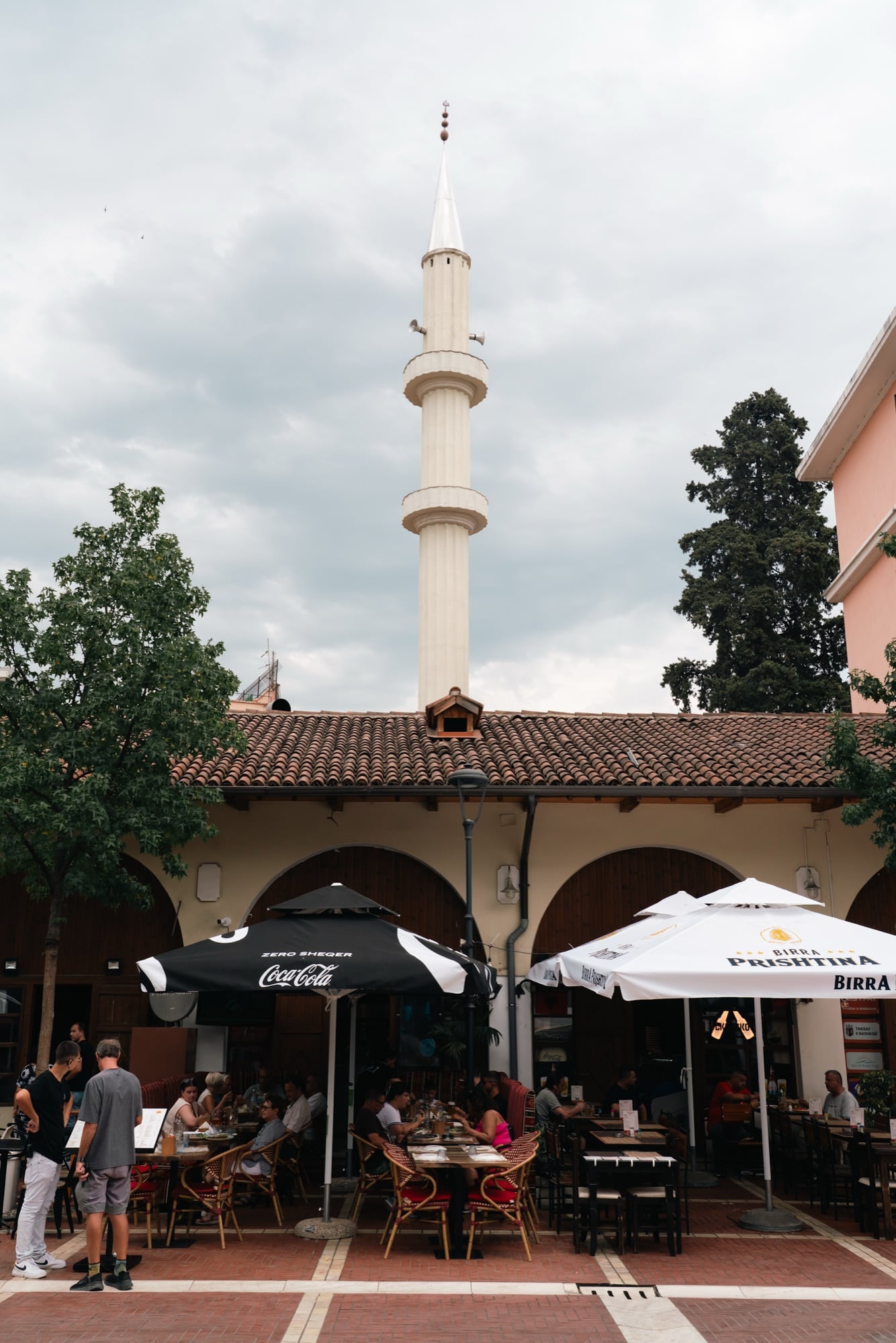 A slender white minaret rising above a tiled-roof arcade and café umbrellas on a busy Tirana square