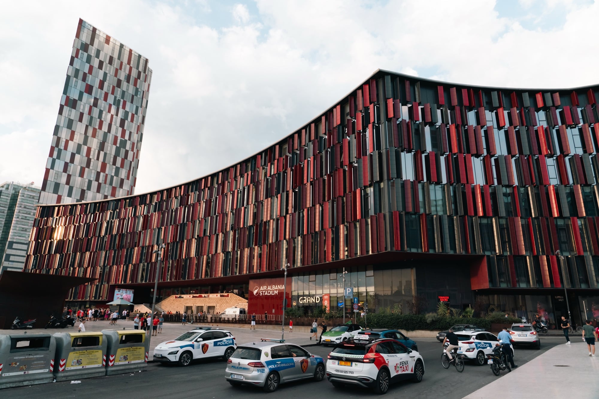 The red-and-black louvered facade of Air Albania Stadium curving beside its checkerboard tower, police cars and taxis in the plaza