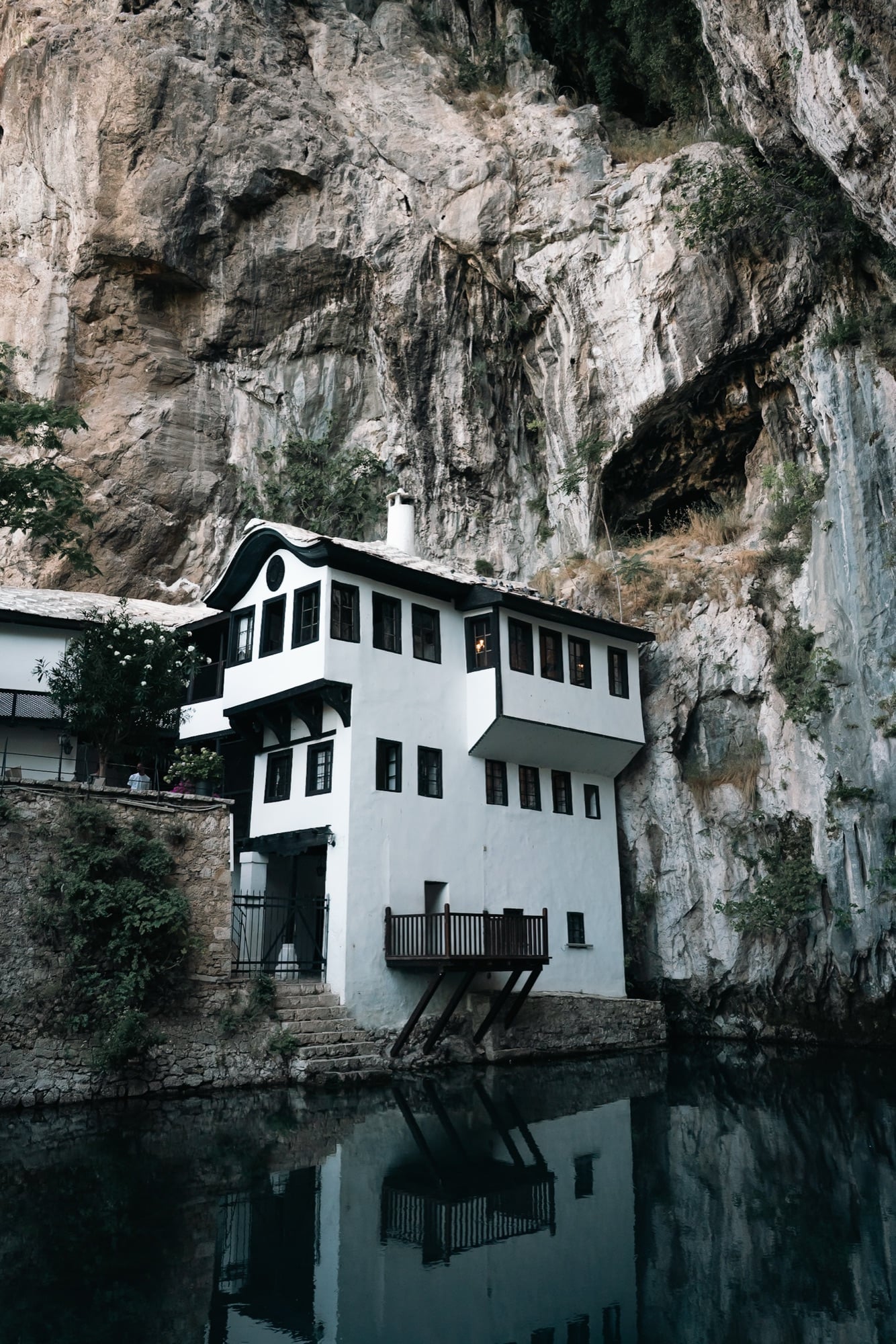 A close view of the black-trimmed white Blagaj Tekija rising from the still emerald pool of the Buna spring, the dark cave mouth gaping in the cliff behind