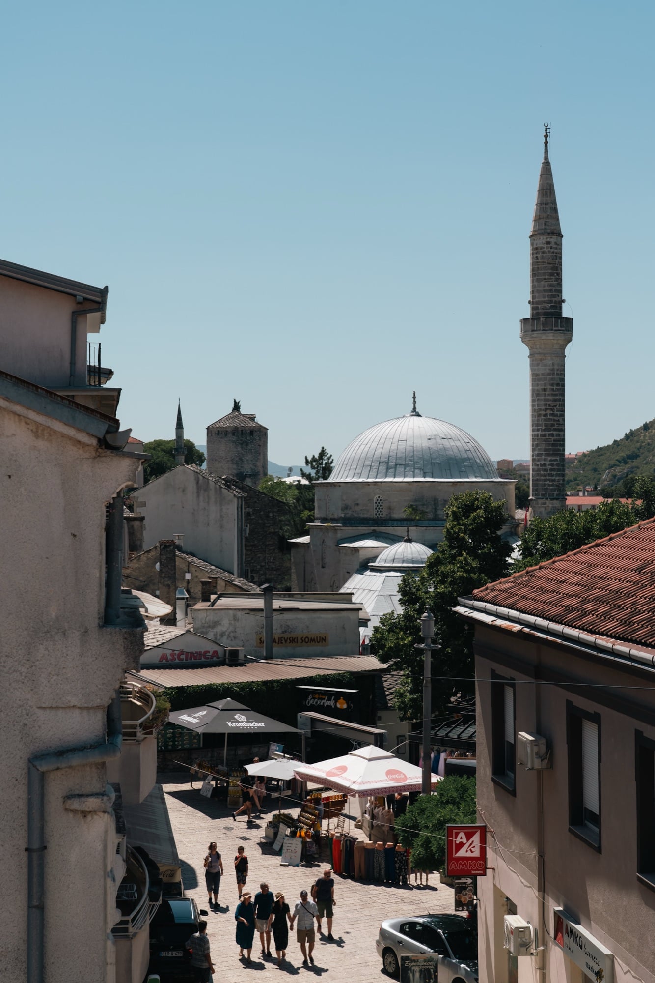 Looking down a Mostar old-town lane past market stalls to the lead dome and slender minaret of the Koski Mehmed-Pasha Mosque beneath bare hills