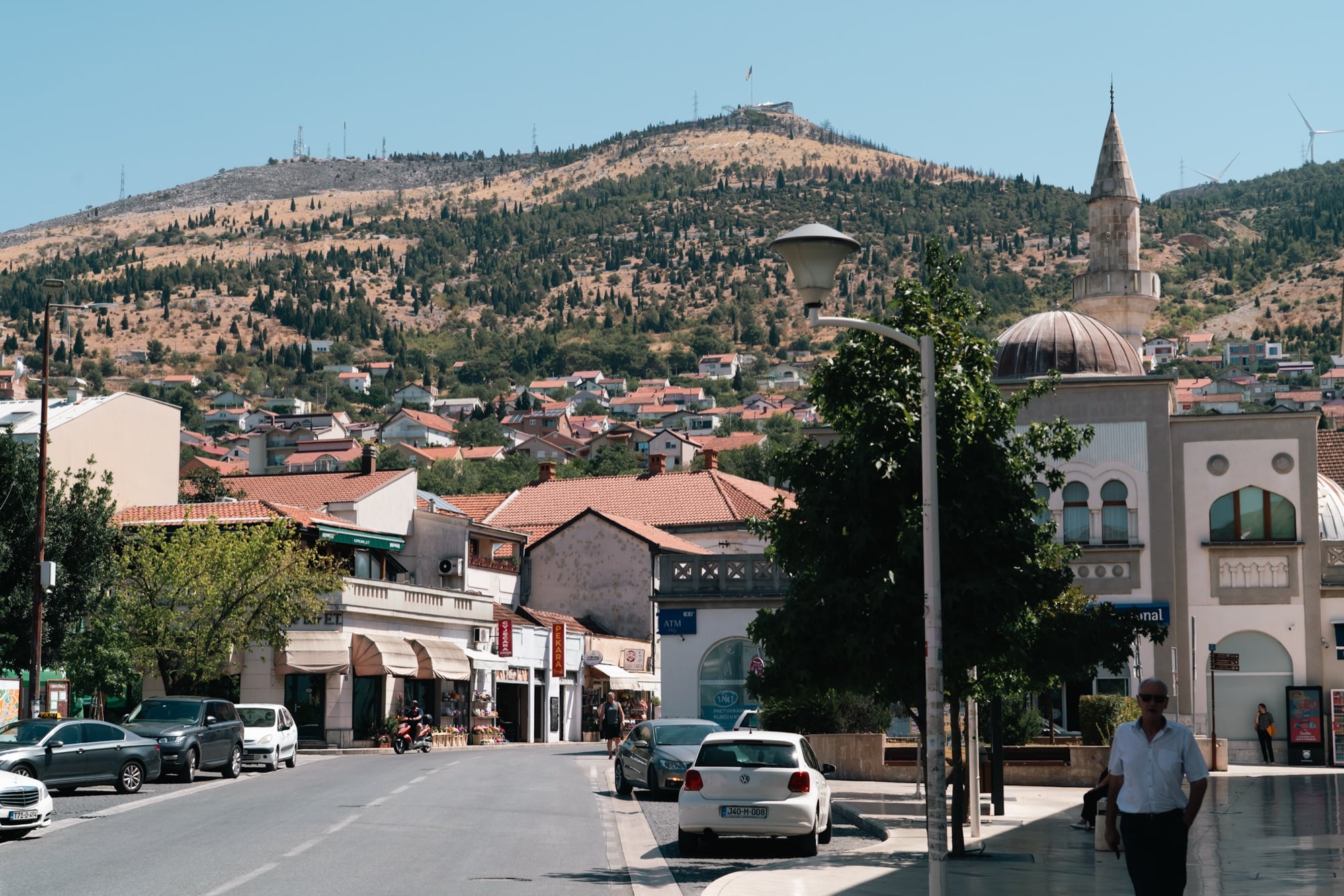 A Mostar street running toward a domed mosque and minaret, red-roofed houses climbing the tawny hillside to a flag on the ridge above