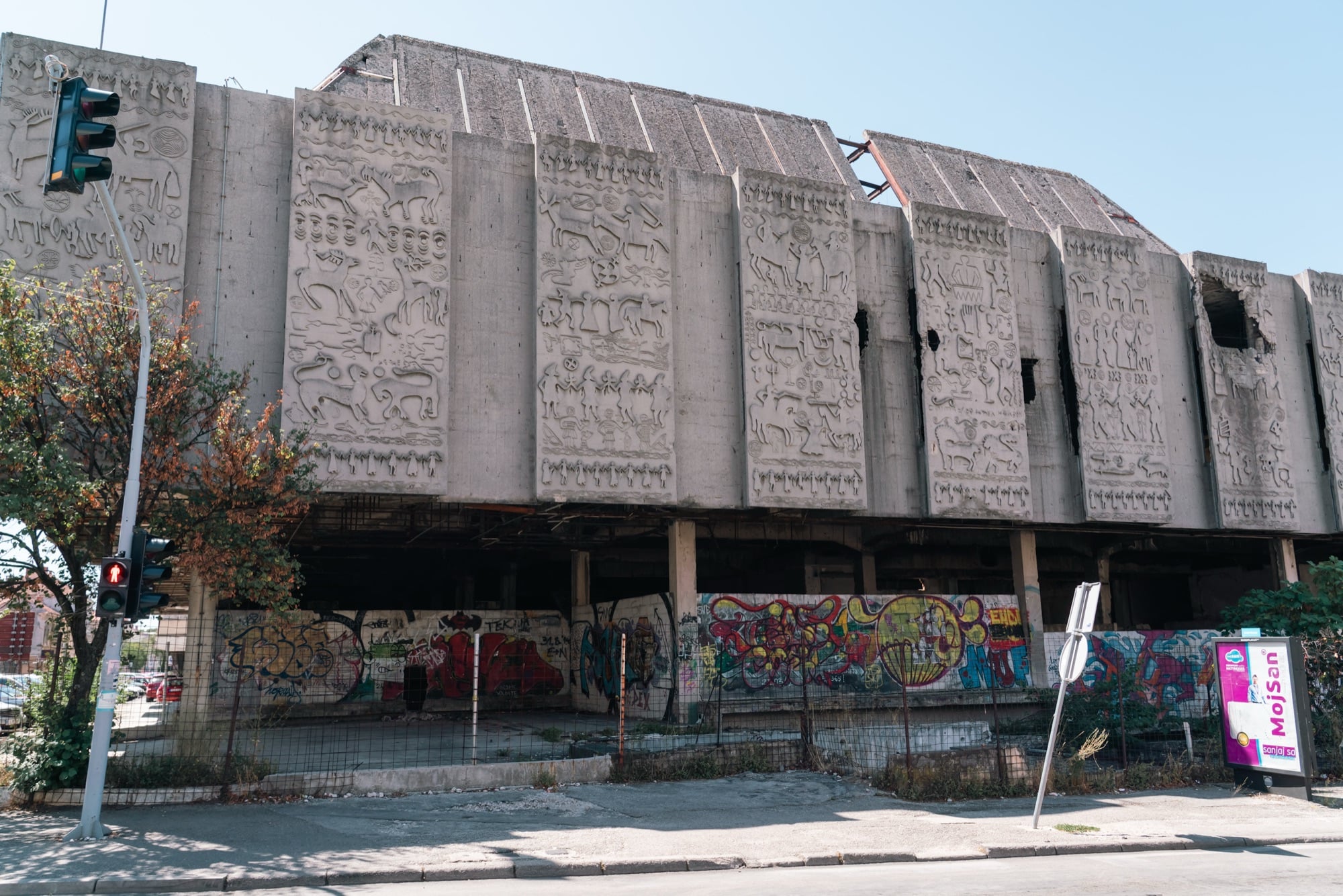 A war-scarred modernist concrete building in Mostar, its façade panels carved with primitive animal and human reliefs, graffiti covering the boarded-up ground floor