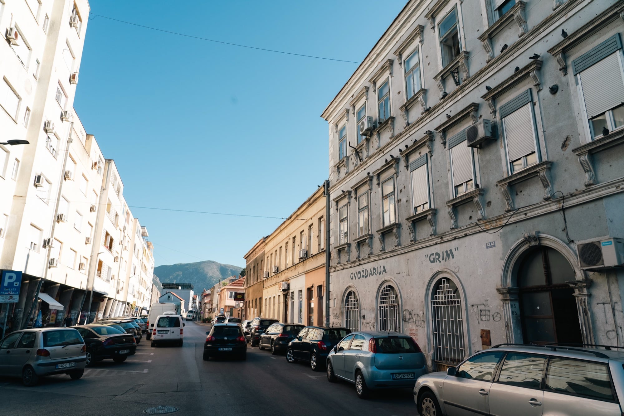 A quiet Mostar street lined with parked cars and bullet-pocked façades, an old 'Gvožđarija Grim' ironmonger's on the right and a bare mountain closing the view