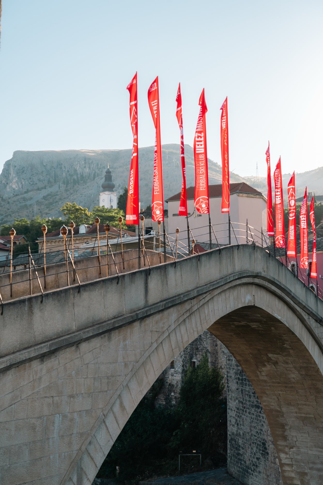 Mostar's Stari Most lined with red Velež football-club flags in the low evening sun, a church tower and rocky cliff rising beyond the arch
