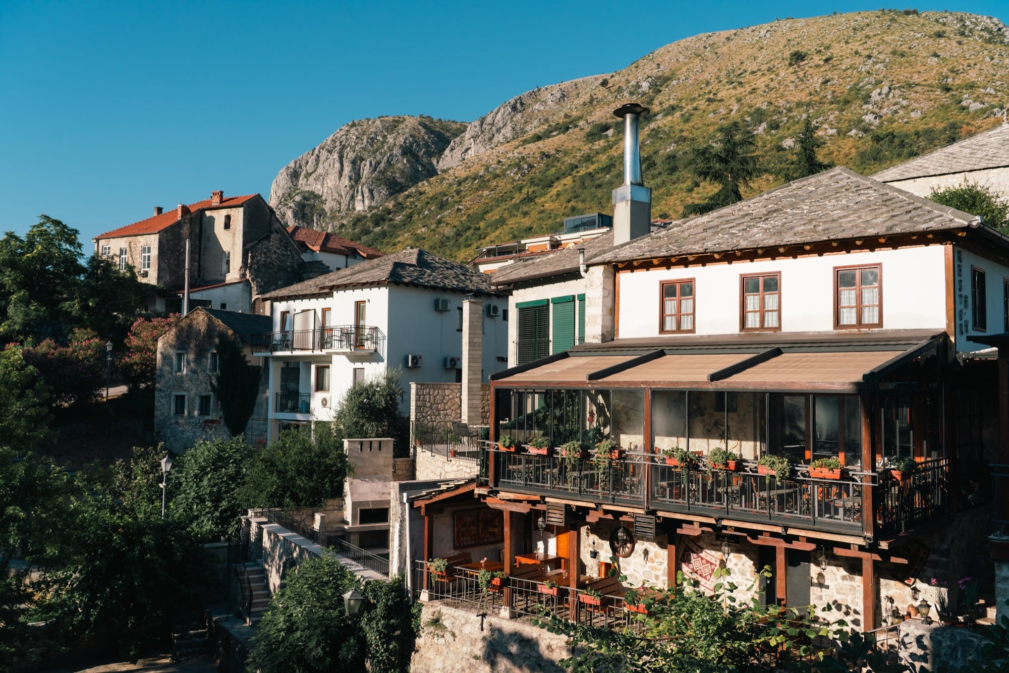 Stone-and-white old-town houses and a glazed restaurant terrace stepping up the Mostar hillside beneath a rugged sunlit mountain