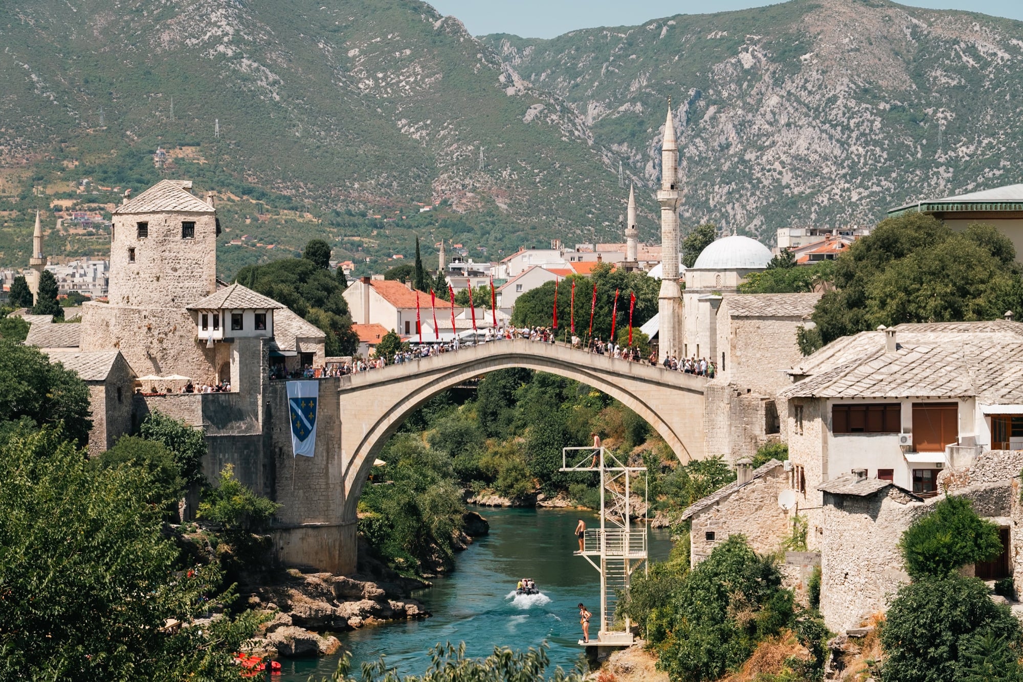The single stone arch of Mostar's Stari Most bridge spanning the turquoise Neretva, crowds and red flags crossing above the diving platform, the bridge's tower and a minaret set against grey mountains