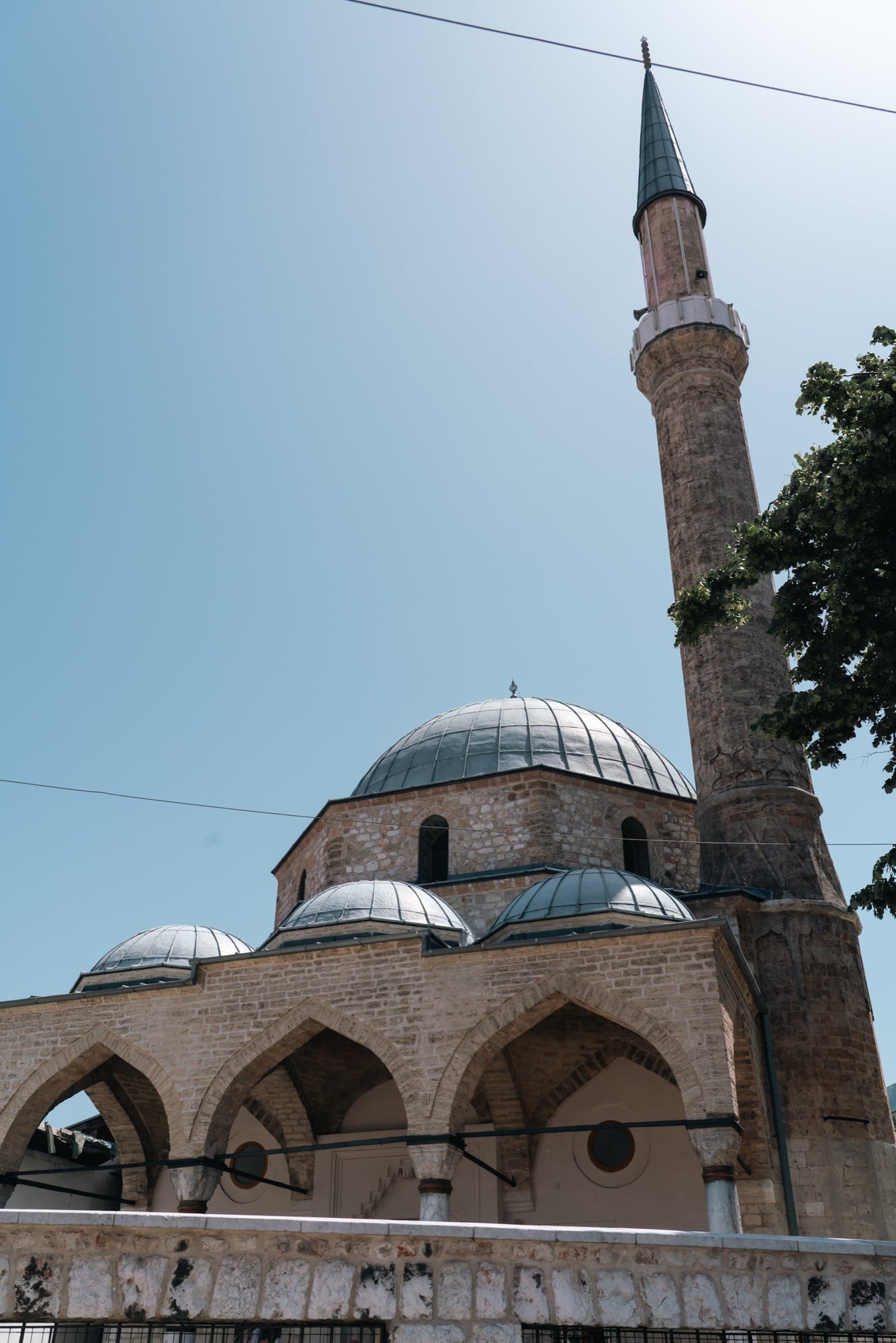 Looking up at the lead dome, arched portico and tall minaret of a stone Ottoman mosque in Sarajevo against a clear sky
