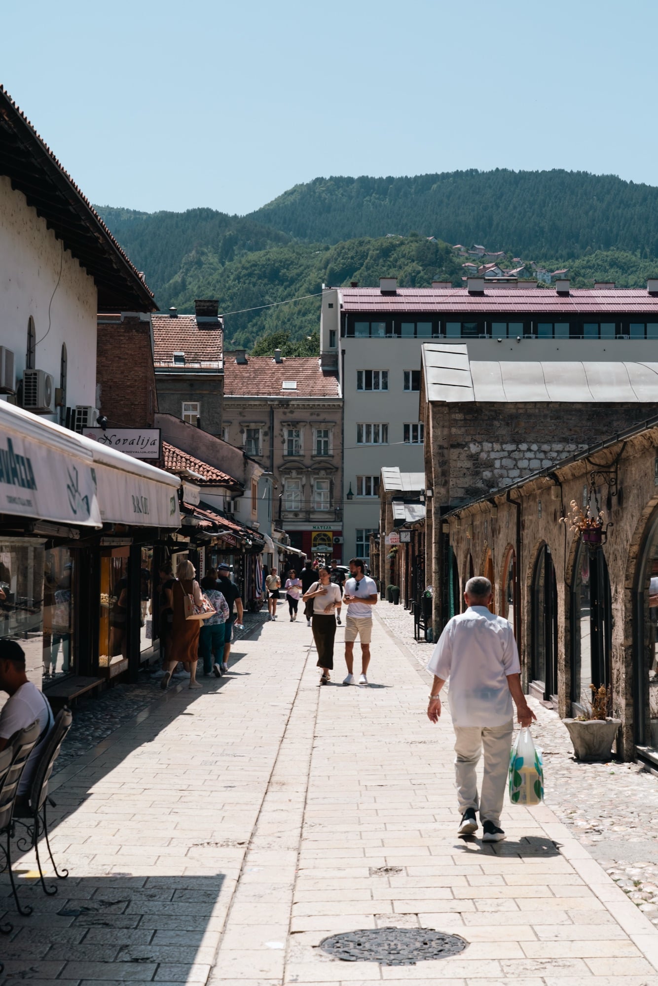 A sunlit Baščaršija shopping street busy with strollers, low stone shopfronts on the right and green mountains closing the view