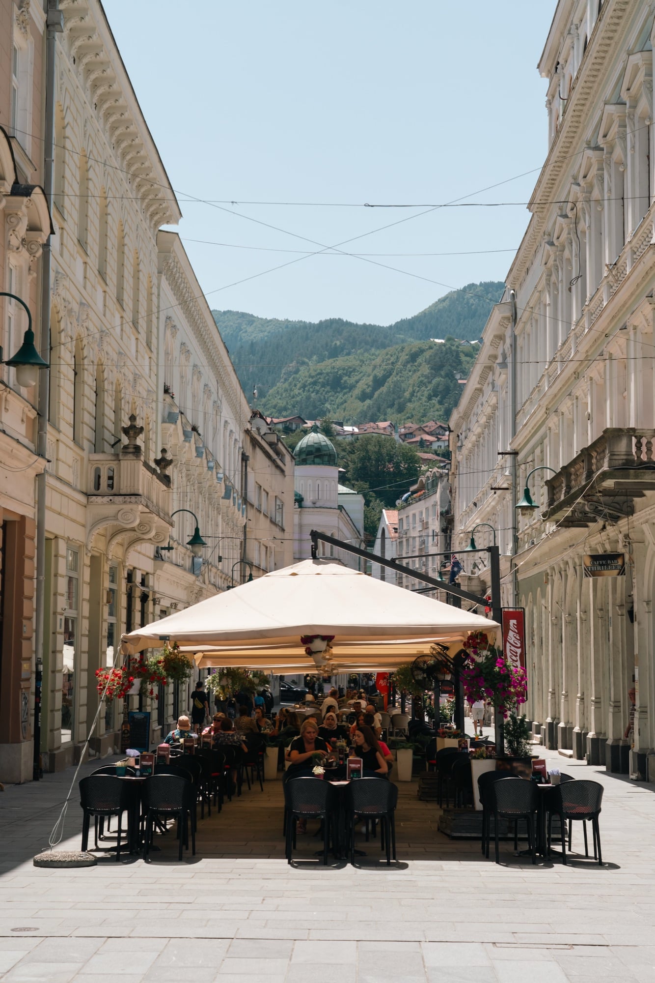 Sarajevo's Ferhadija promenade with a café spread under a broad cream awning between grand pale façades, a green mountain and church dome closing the vista