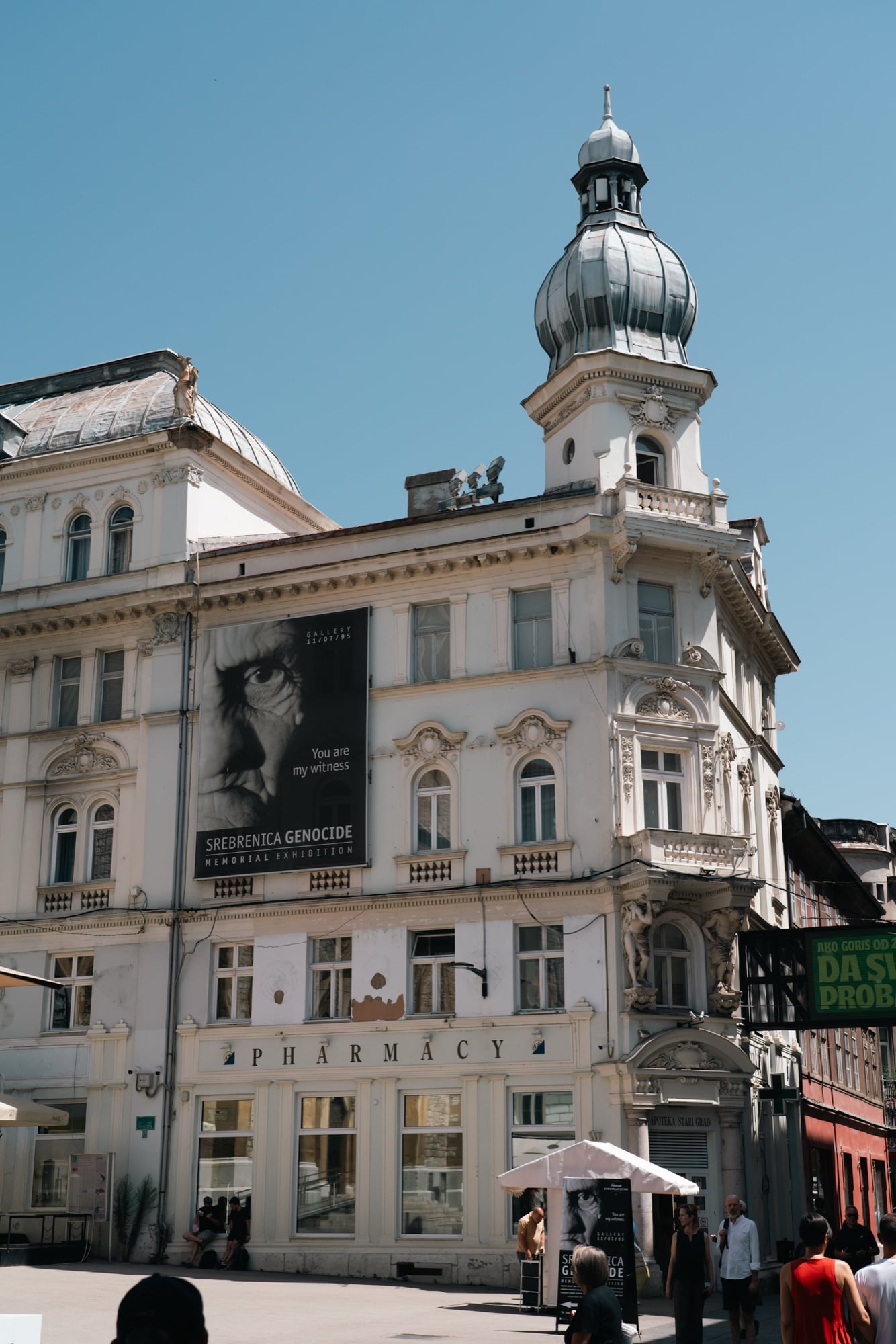 A grand corner building in Sarajevo topped by a silver onion-dome turret, its façade hung with the Srebrenica Genocide memorial banner reading 'You are my witness' above a pharmacy