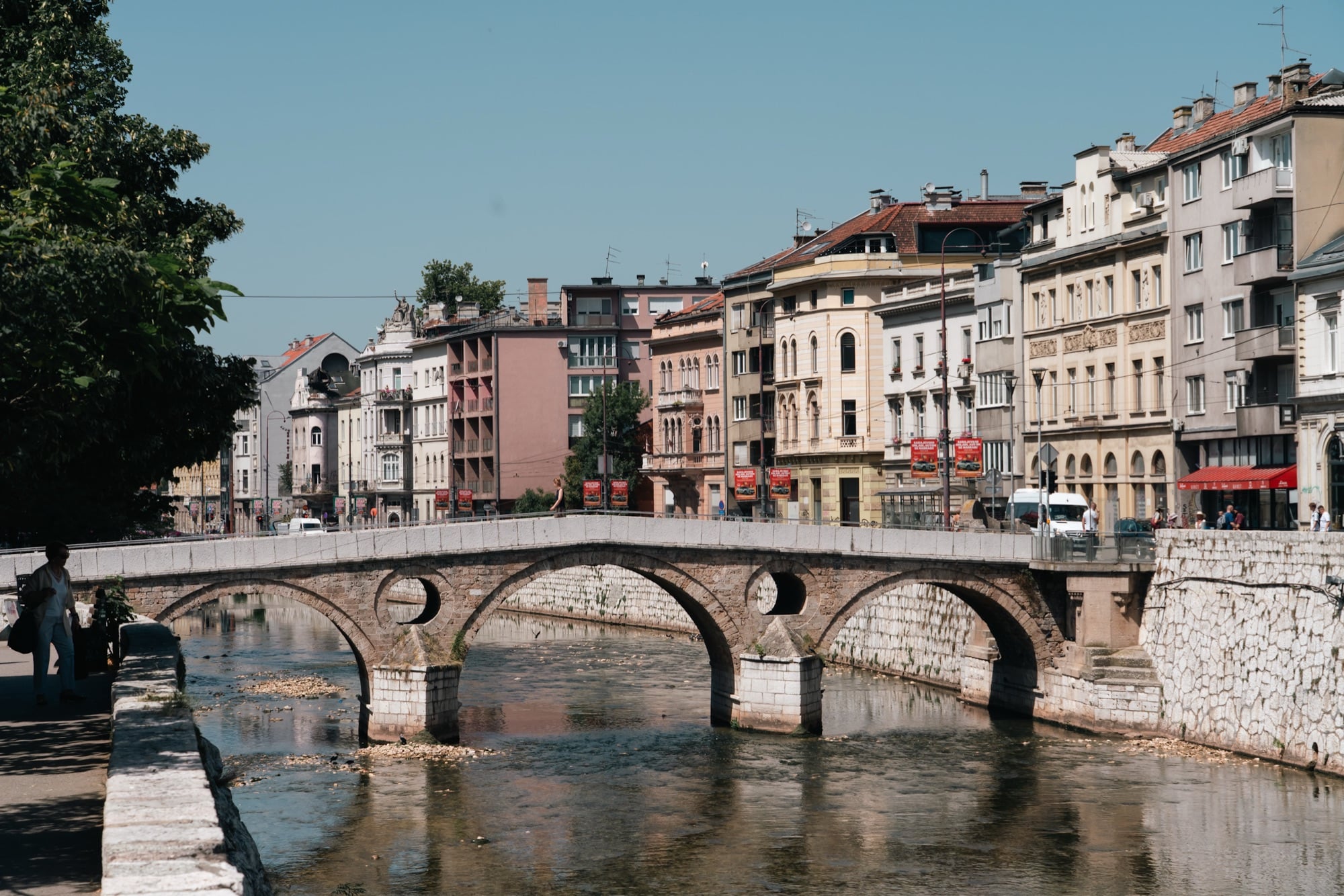 The stone arches of the Latin Bridge spanning the shallow Miljacka, a row of pastel Sarajevo façades lining the far embankment