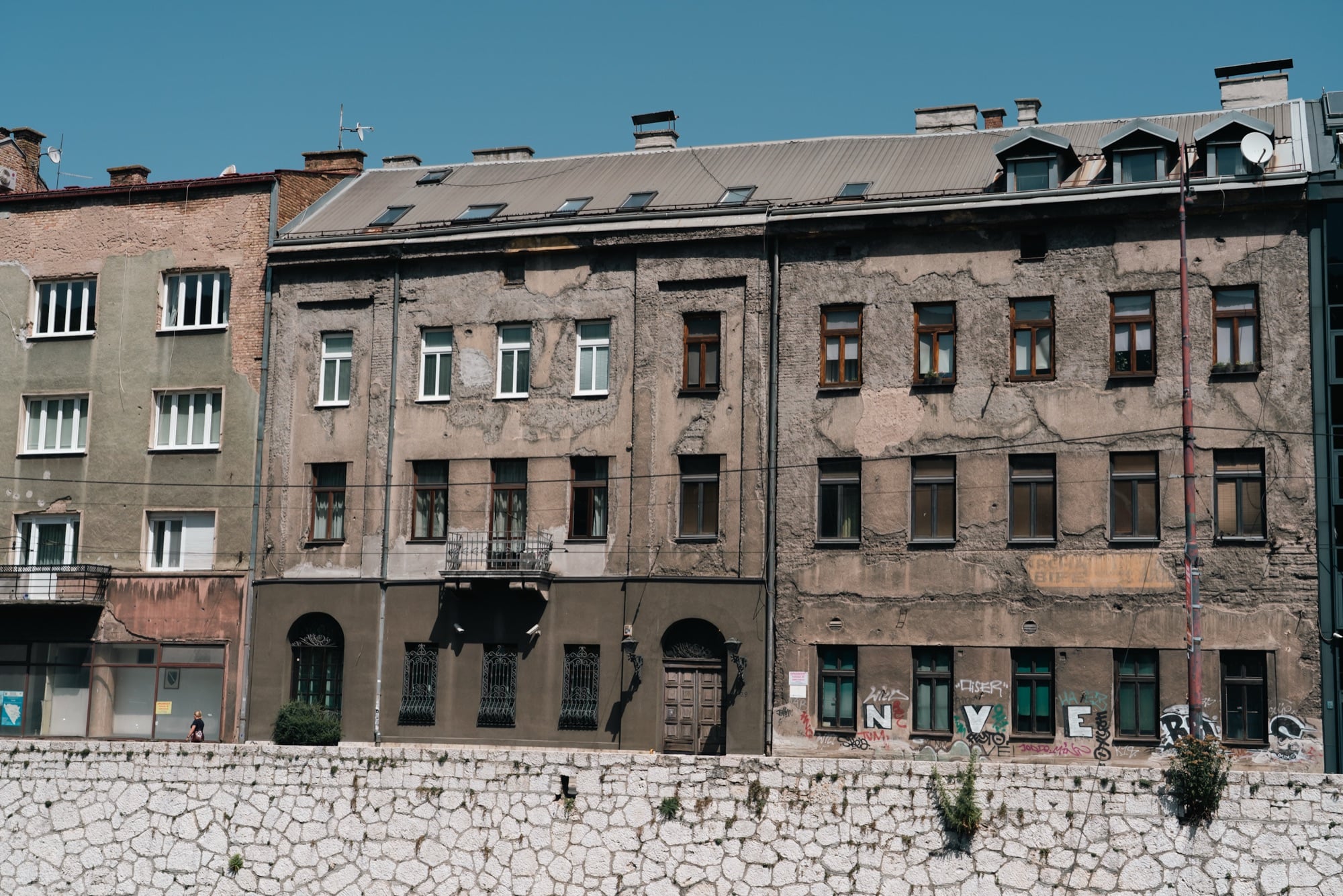 Weathered, war-marked apartment façades rising above the white stone embankment of the Miljacka, one block crumbling beside a repainted neighbour