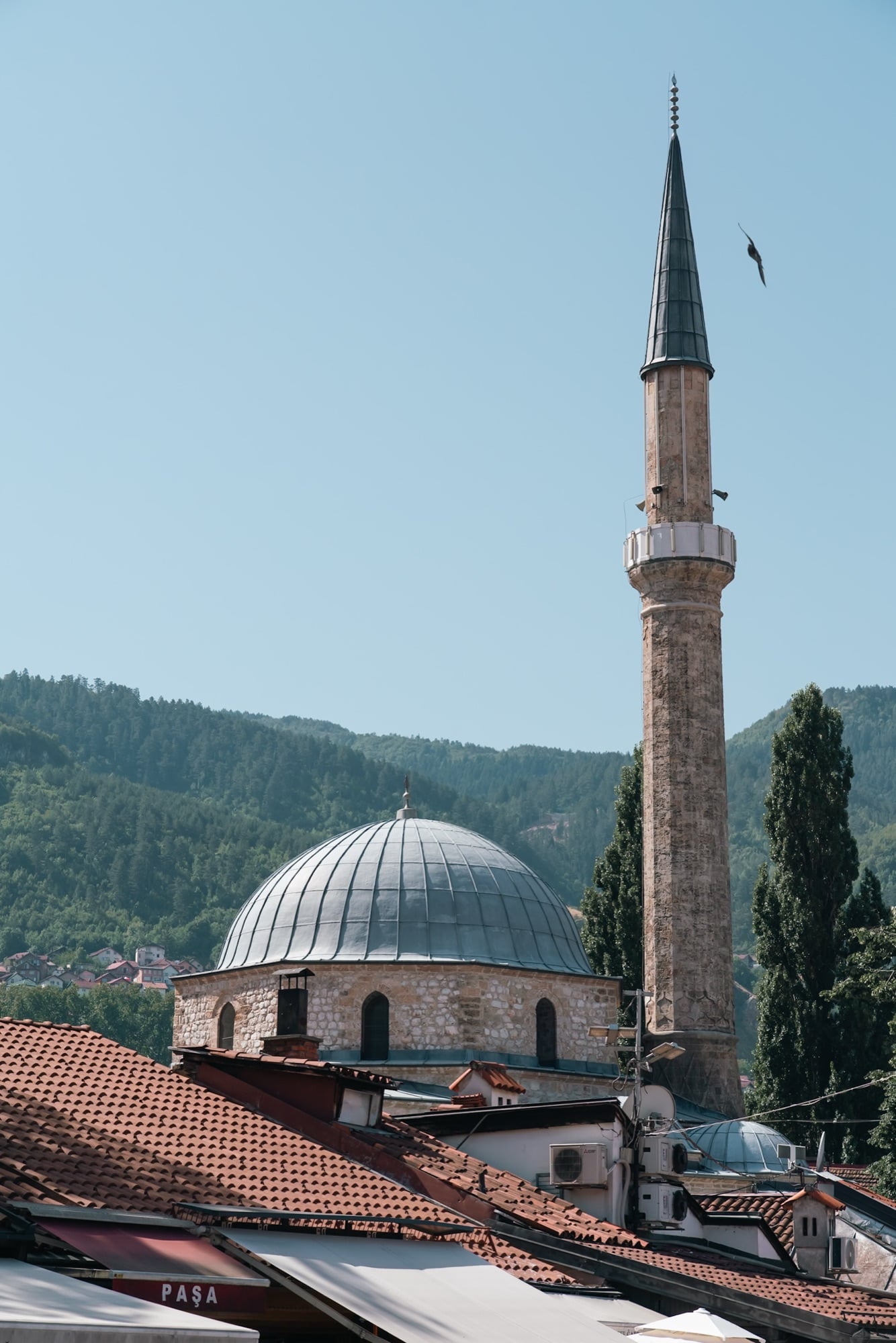 The lead dome and towering stone minaret of the Gazi Husrev-beg Mosque above Baščaršija's terracotta rooftops, a bird wheeling past the wooded hills