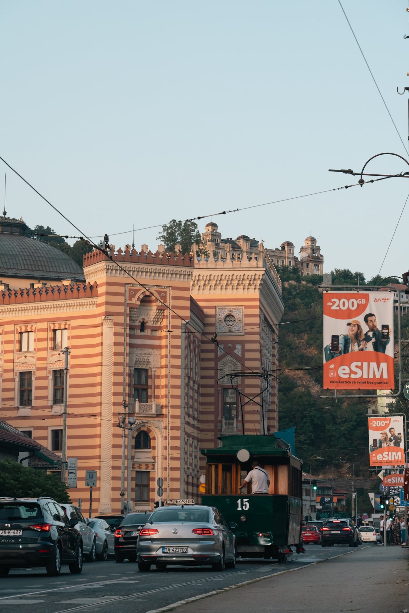 A green heritage tram number 15 rolling past the red-and-cream striped corner of Sarajevo's City Hall at dusk, a hilltop villa above the traffic