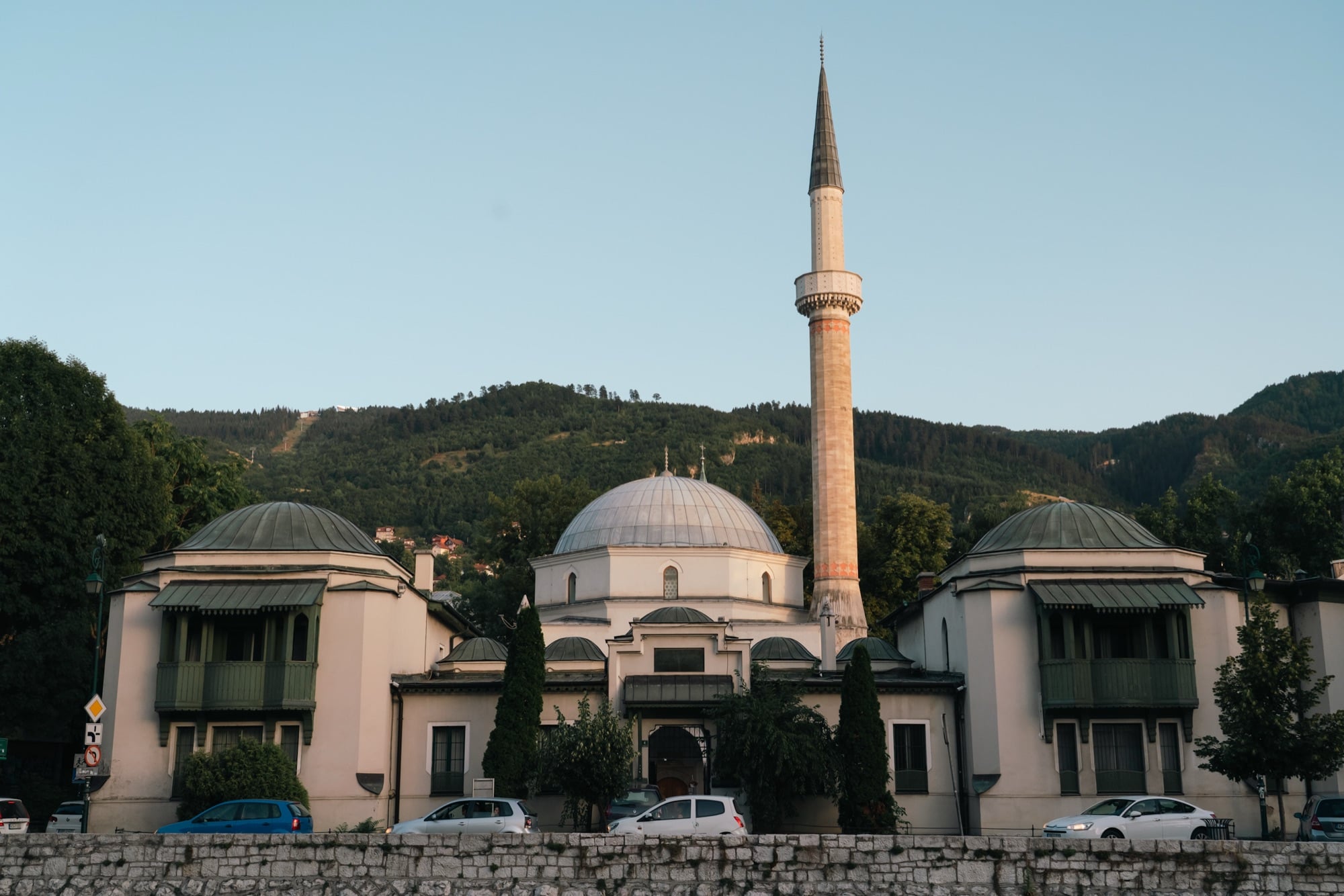 The domed Emperor's Mosque and its tall stone minaret beside the Miljacka, wooded hills rising behind and cars parked along the embankment