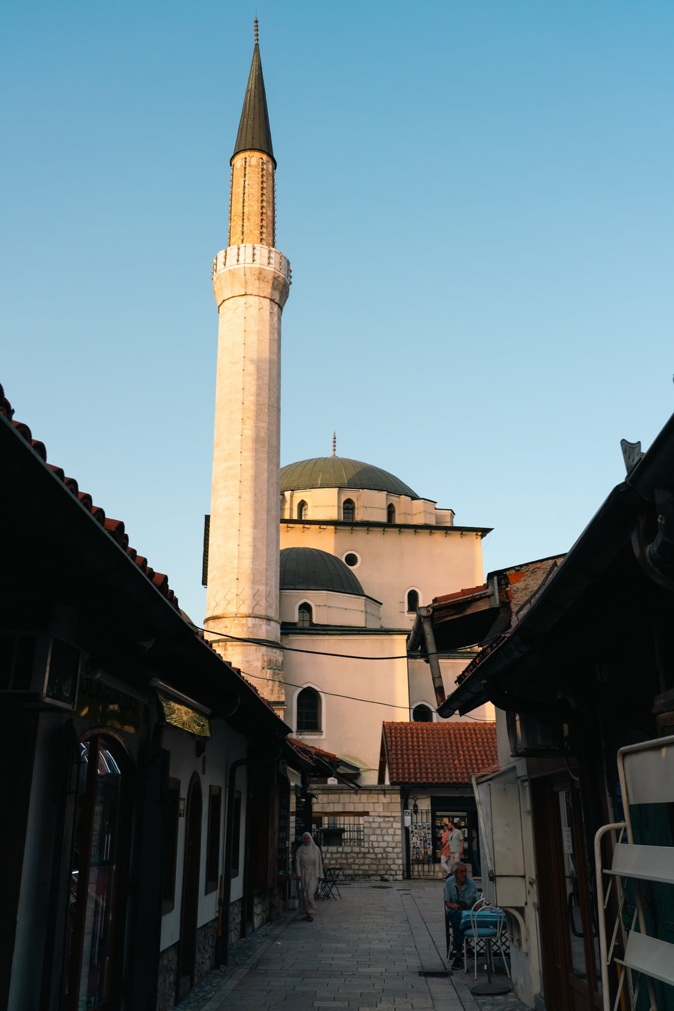 A Baščaršija lane at golden hour framing the sunlit minaret and lead domes of a mosque, café chairs and shopfronts lining the cobbles