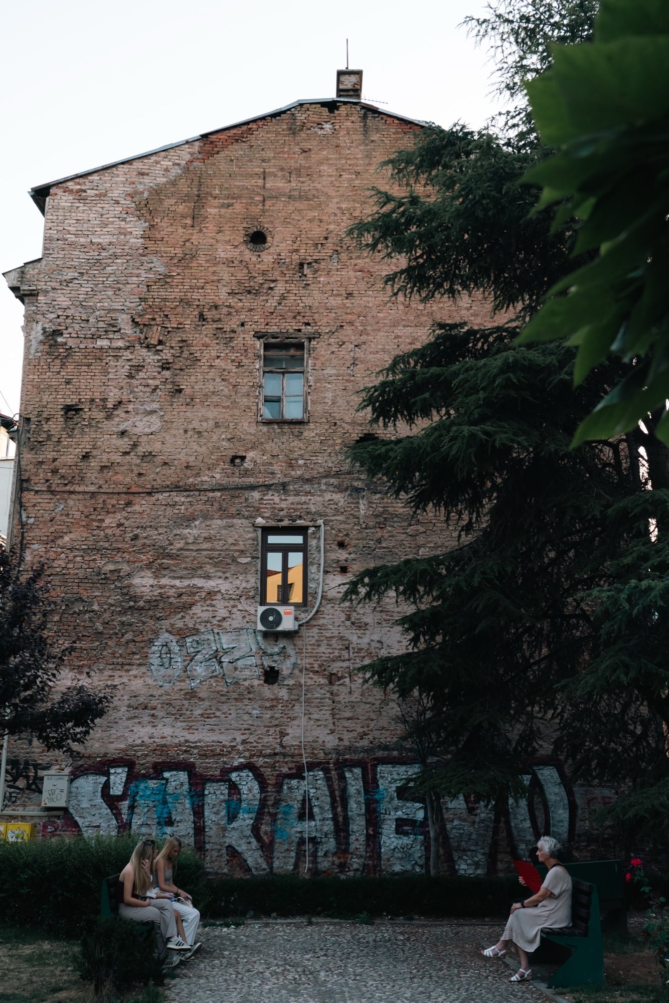 A towering war-scarred brick gable wall tagged with 'SARAJEVO' in a small Baščaršija park, people resting on benches in the fading light