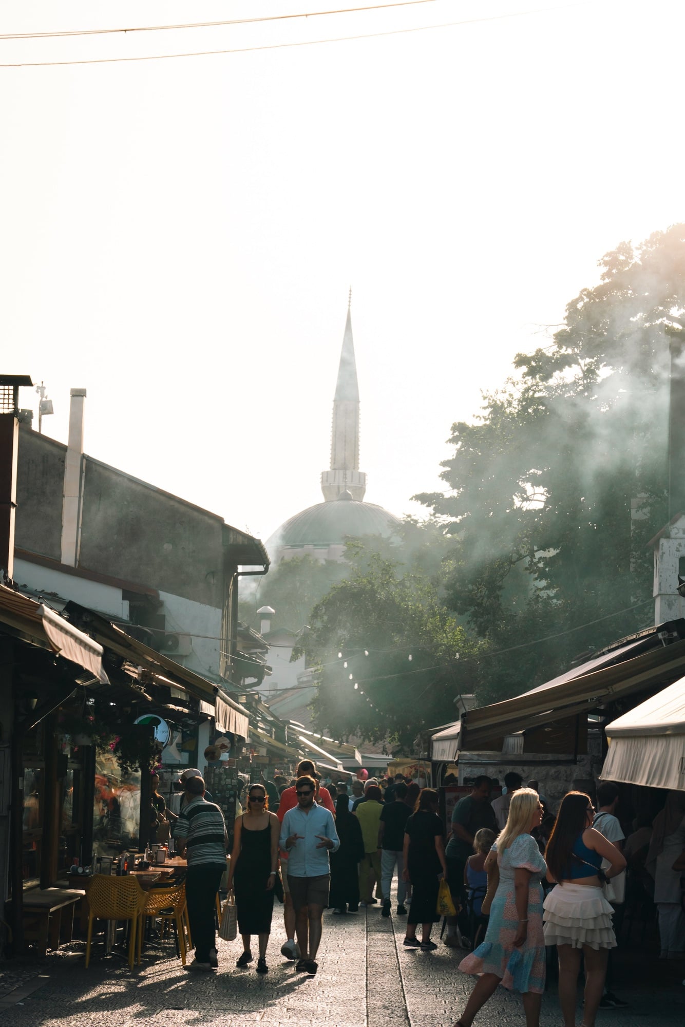 Crowds filling a Baščaršija market street in hazy backlit evening sun, the dome and minaret of a mosque silhouetted through drifting smoke