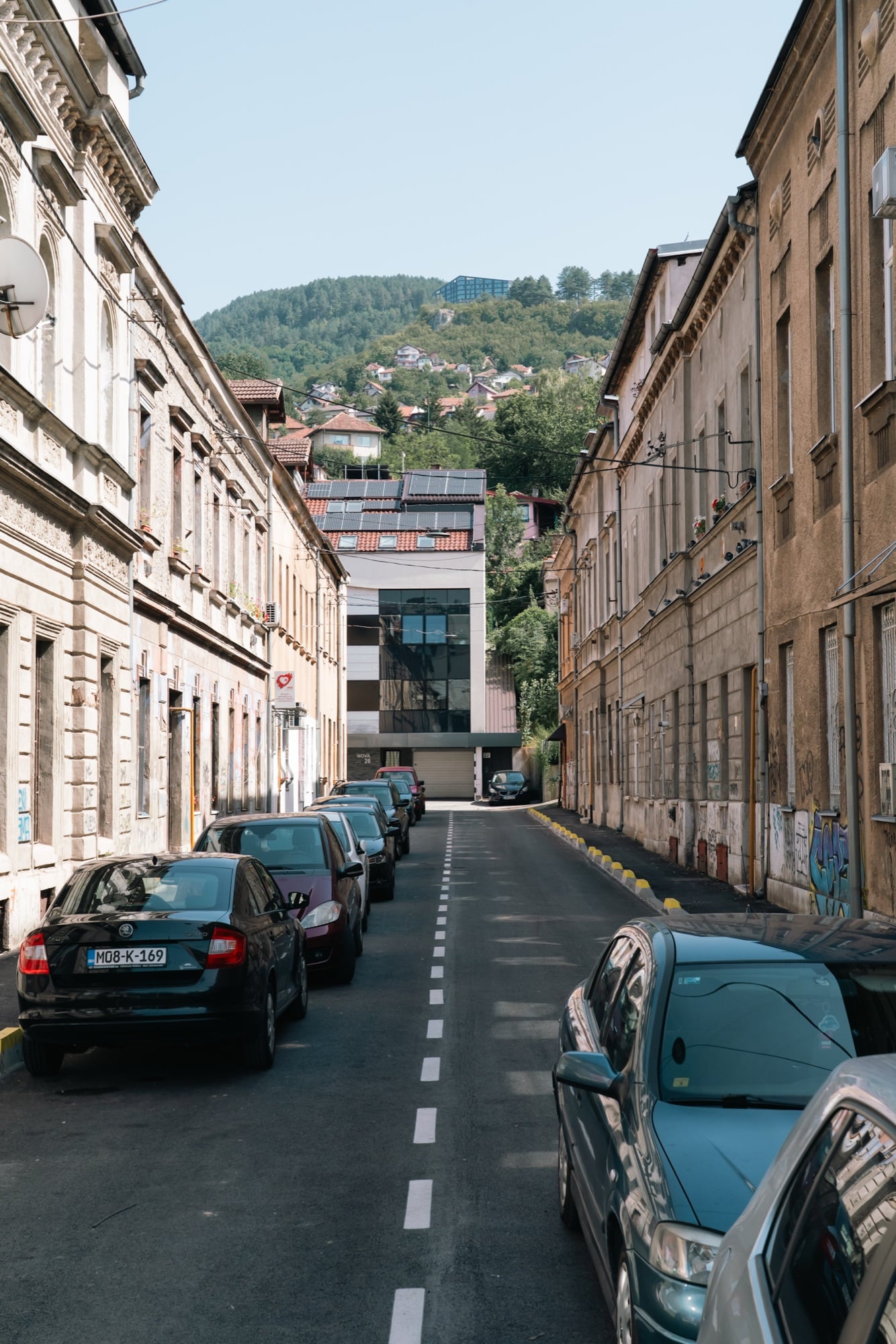 A steep narrow Sarajevo side street of parked cars and old façades, wooded slopes and a hilltop building rising at its head