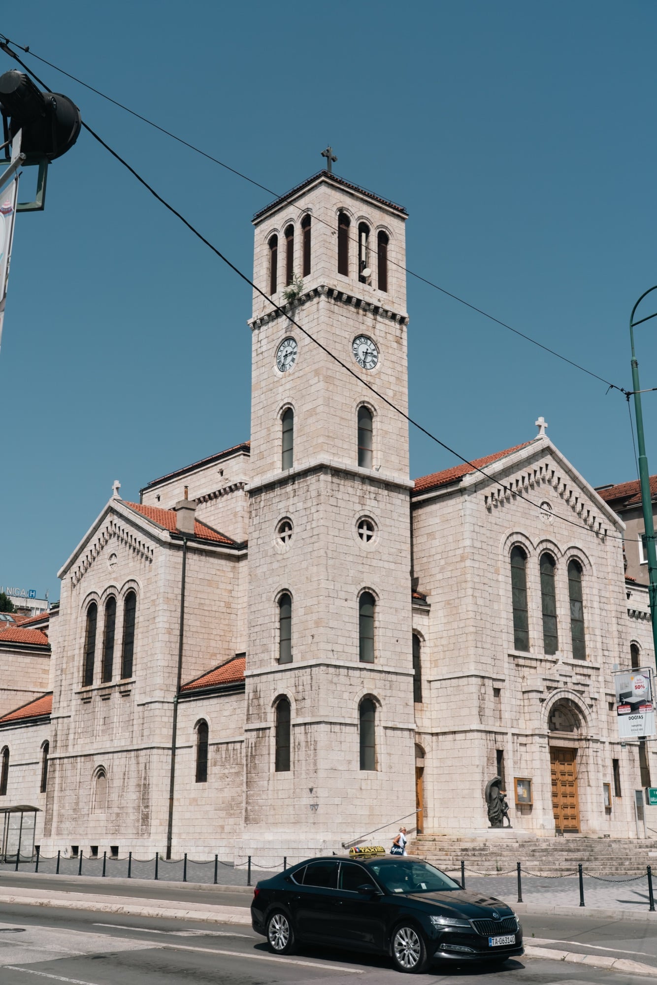 The stone Romanesque bell tower and clock of St Joseph's Church in Sarajevo, a black taxi passing on the road below a clear blue sky