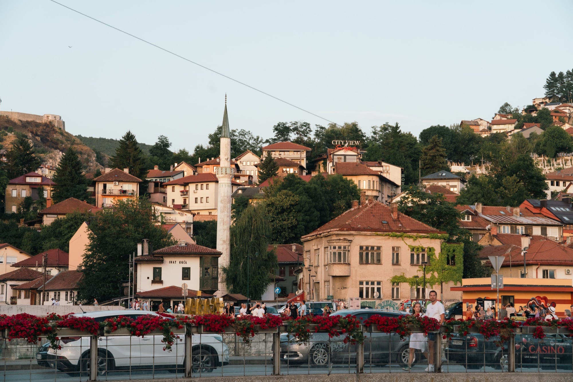 Sarajevo's old-town houses and a white minaret climbing the hillside at dusk, the riverside 'Inat Kuća' below and red flowers spilling from a bridge railing