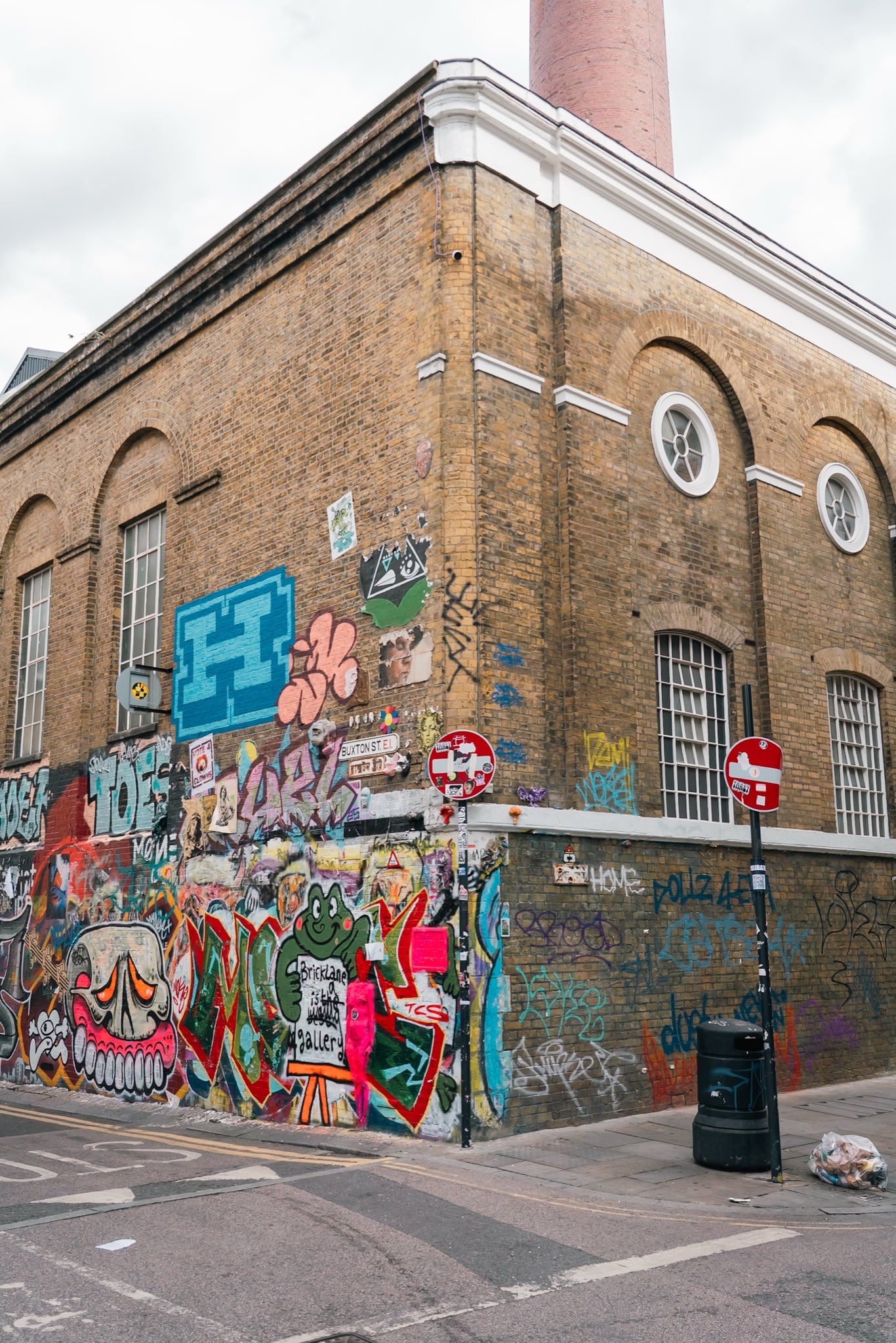 A graffiti-covered brick corner building beneath a tall chimney off Brick Lane