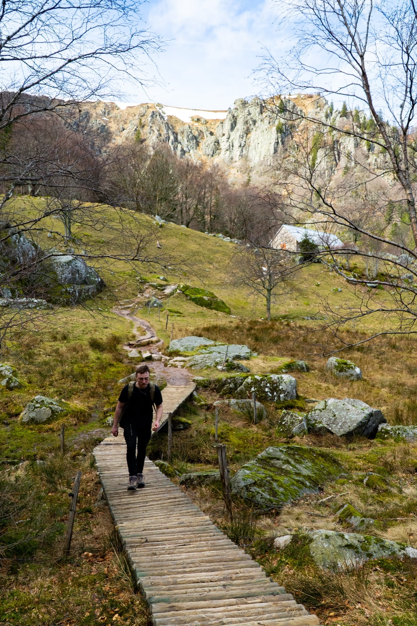 A hiker on a wooden boardwalk trail climbing through moss and rock below the Vosges crags near Gérardmer