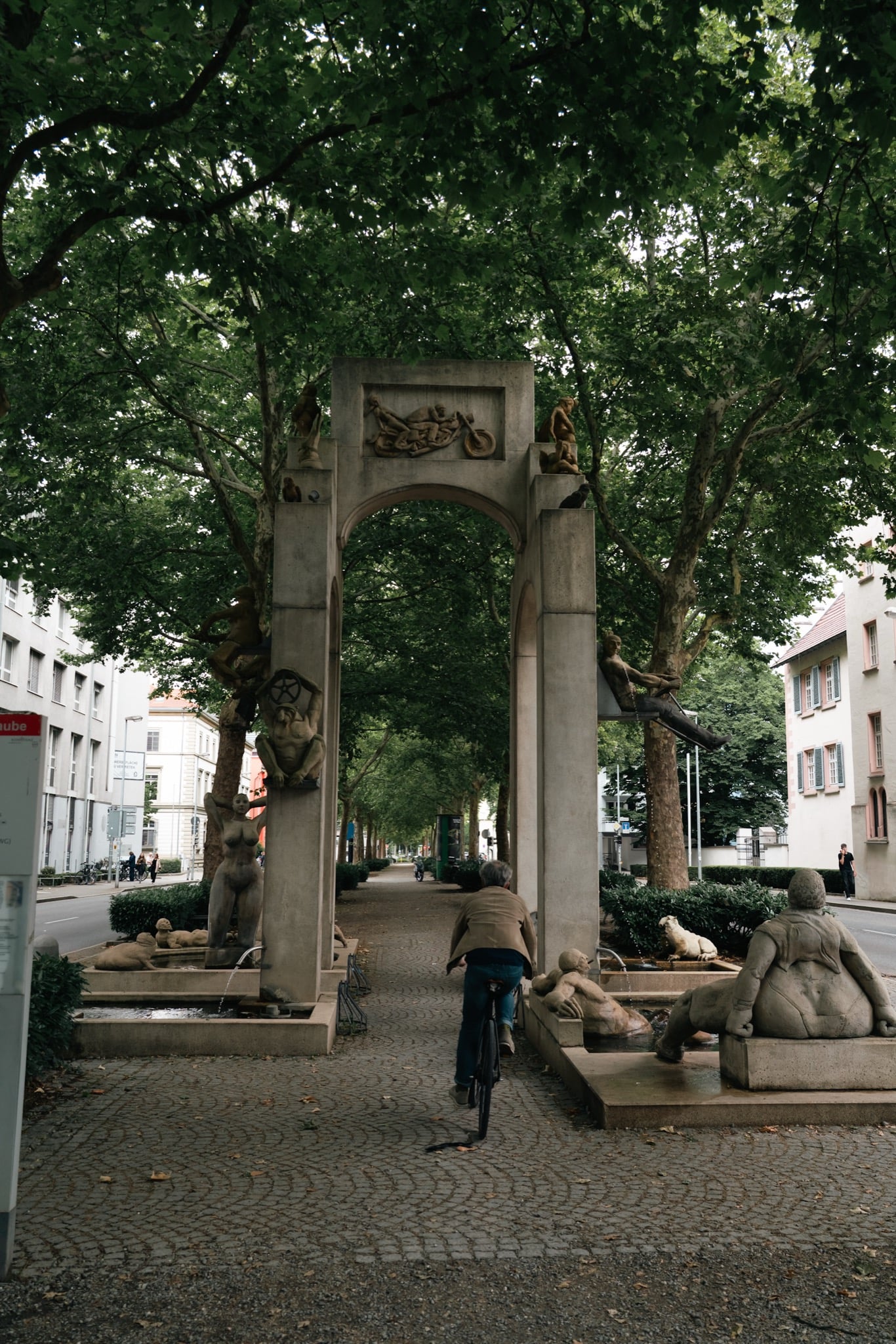 A cyclist passing under a sculpture-lined fountain gateway on a tree-shaded Konstanz avenue