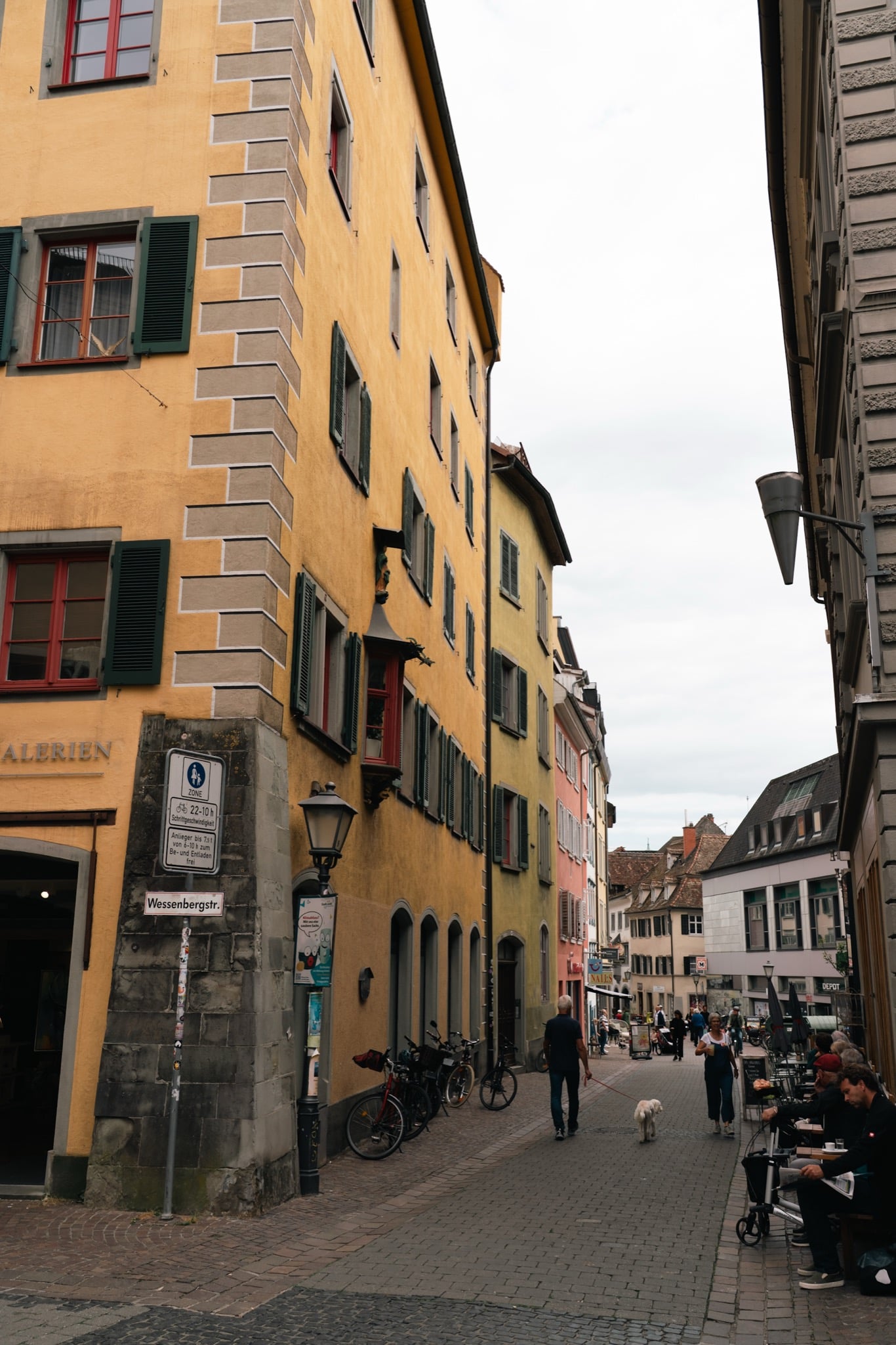 A colourful old-town lane in Konstanz with a dog walker and café tables