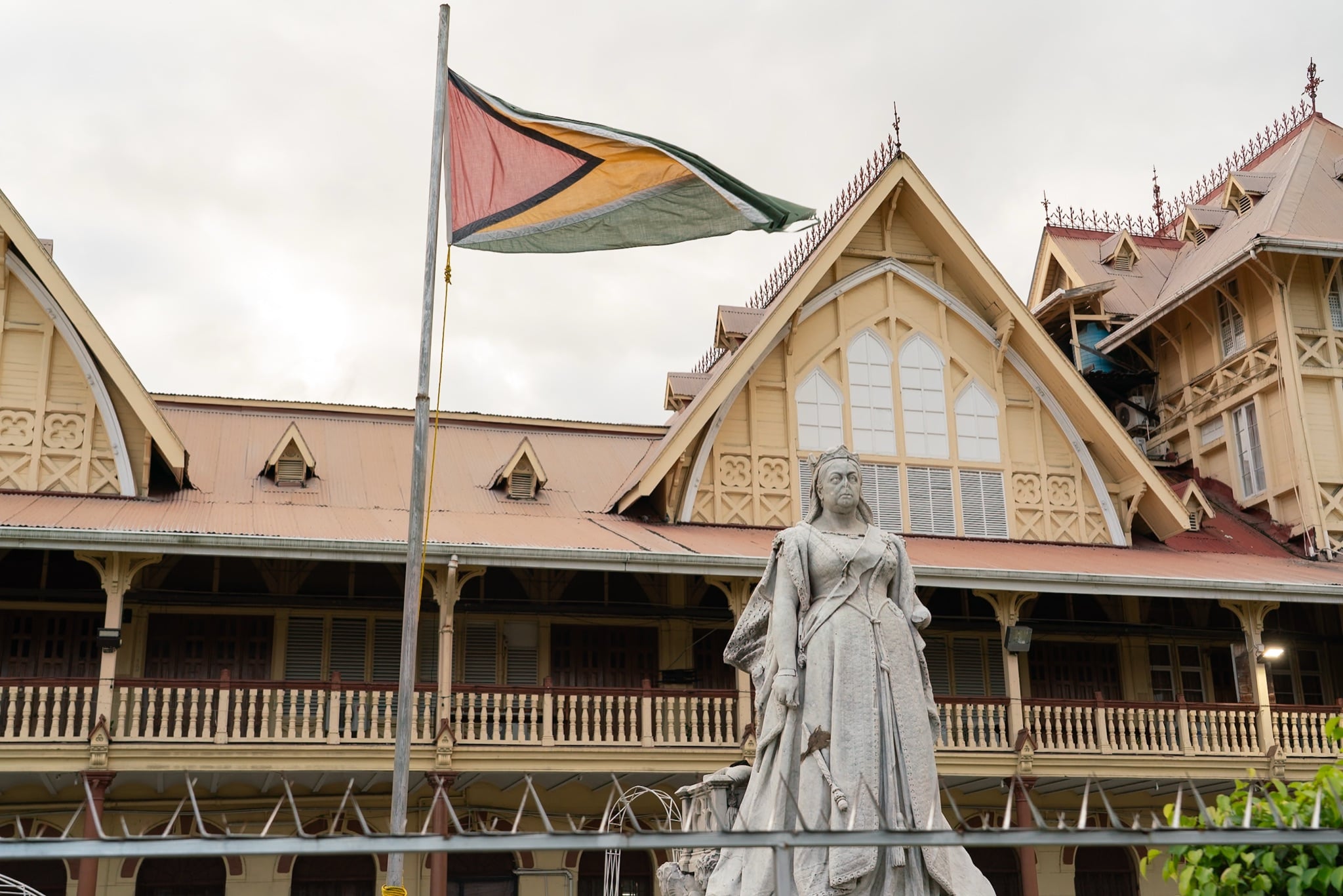 A statue and the Guyana flag before a grand wooden colonial building