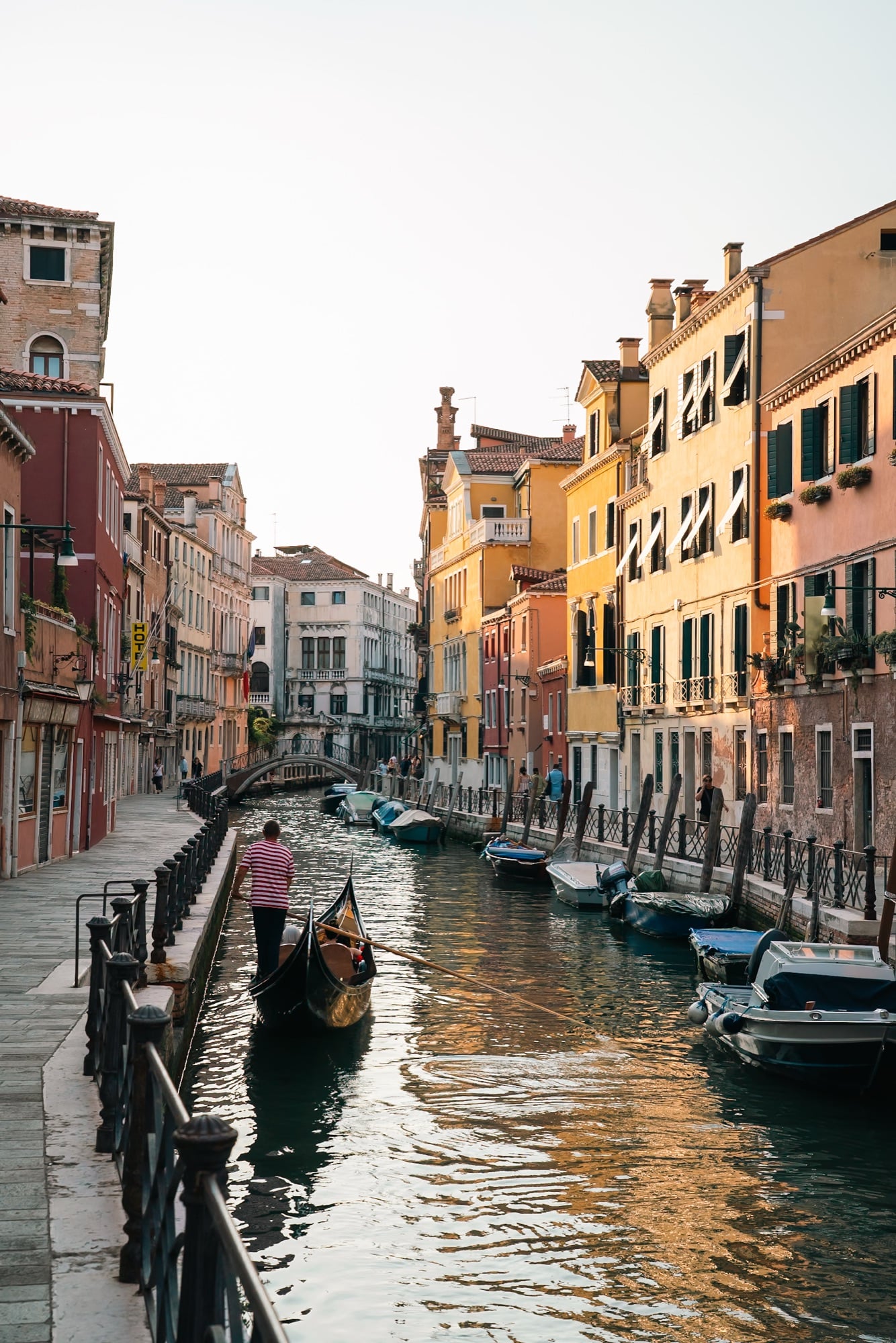 A gondolier steering his gondola along a quiet side canal at golden hour, moored motorboats and a small arched bridge ahead