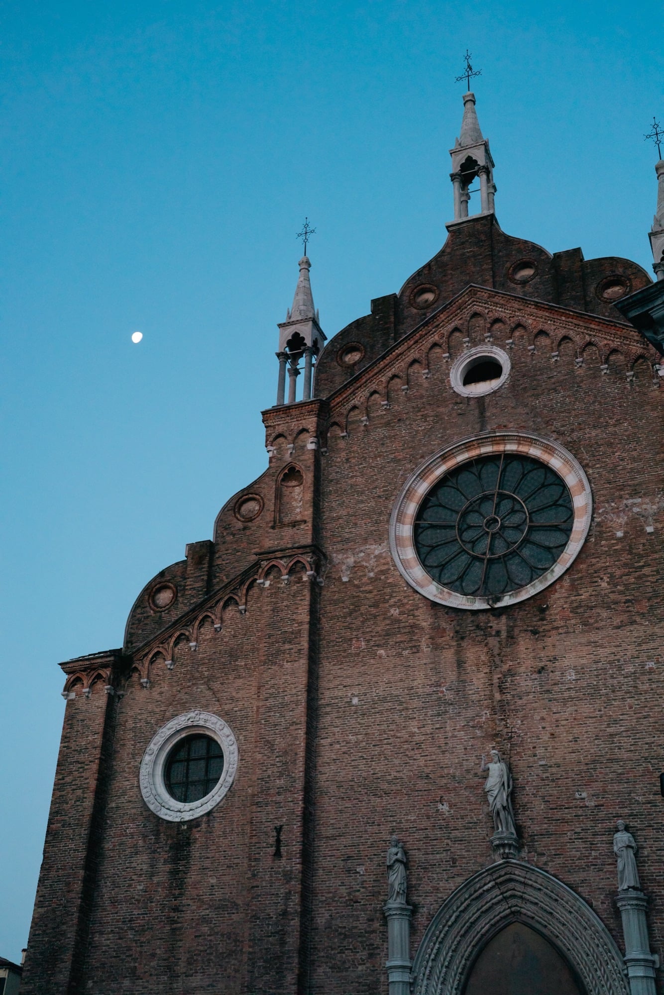 The brick Gothic facade of the Frari basilica with its great rose window and crocketed gables, a rising moon in the dusk sky
