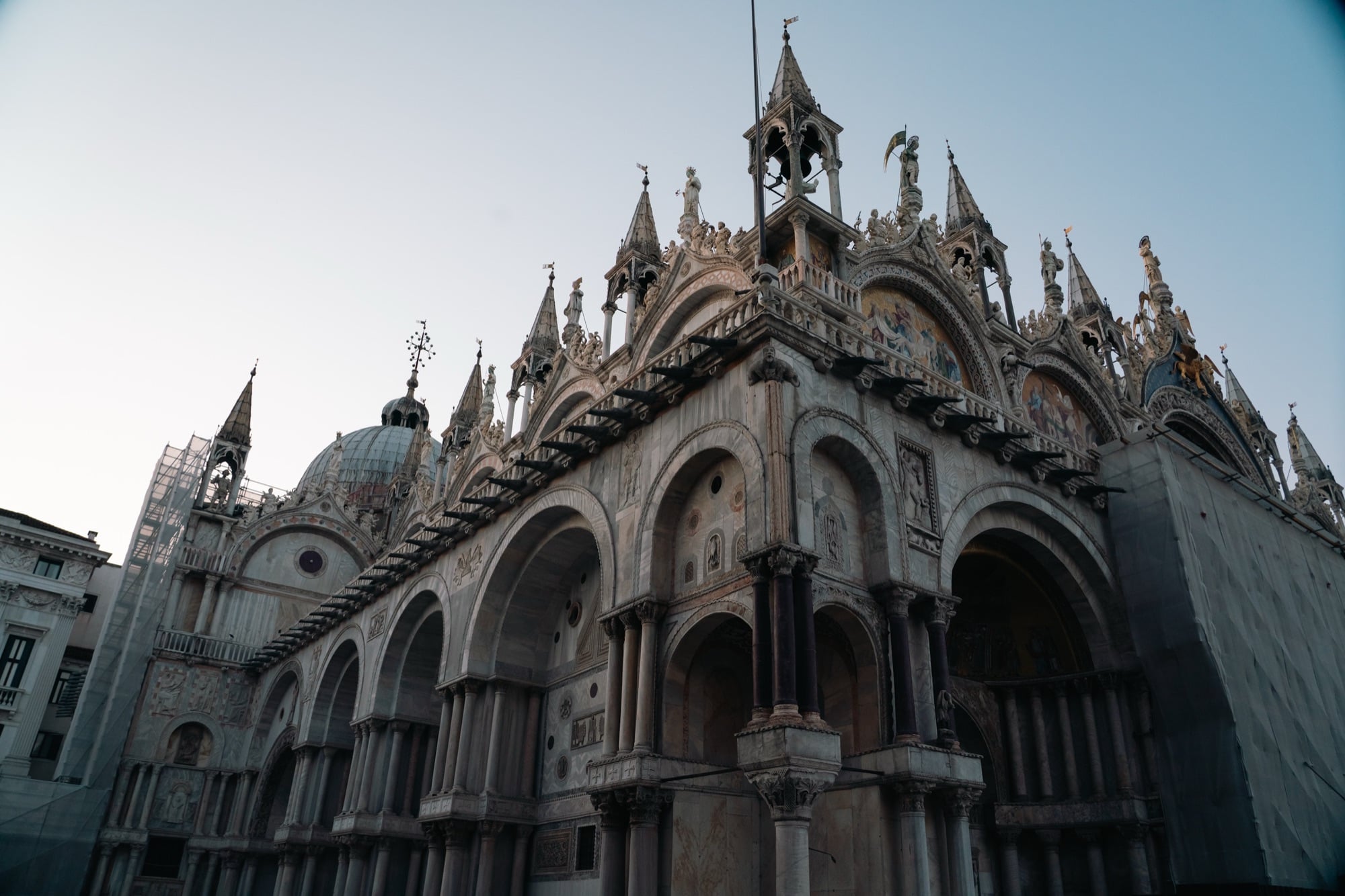The ornate marble facade of St Mark's Basilica at first light, its domes, spires, mosaics and clustered statues catching the pale sky
