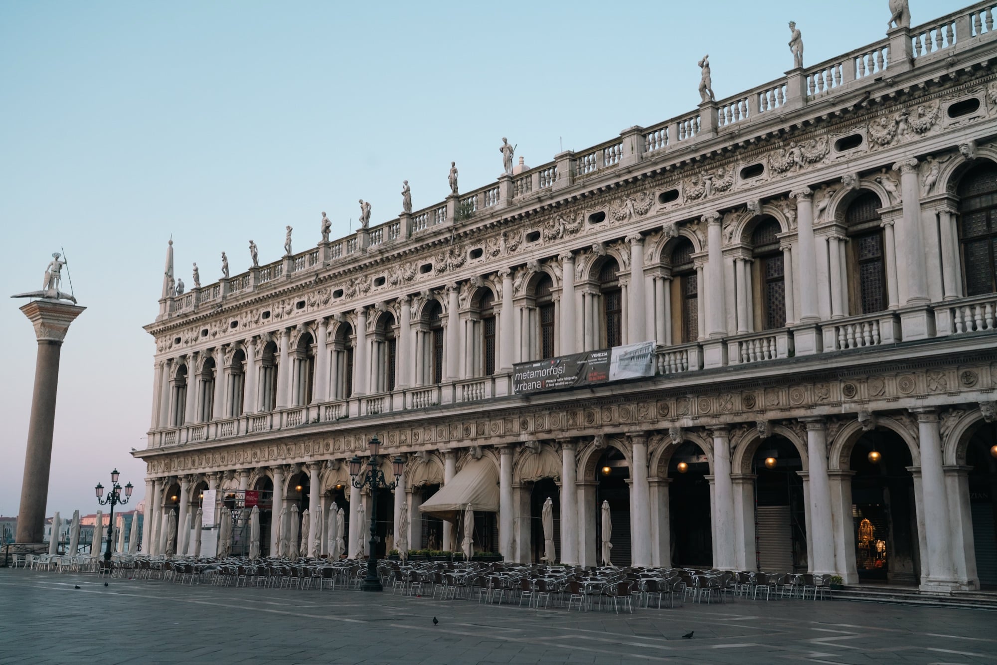 The statue-crowned Renaissance arcade of the Biblioteca Marciana at dawn, the column of Saint Theodore at left and empty café chairs below