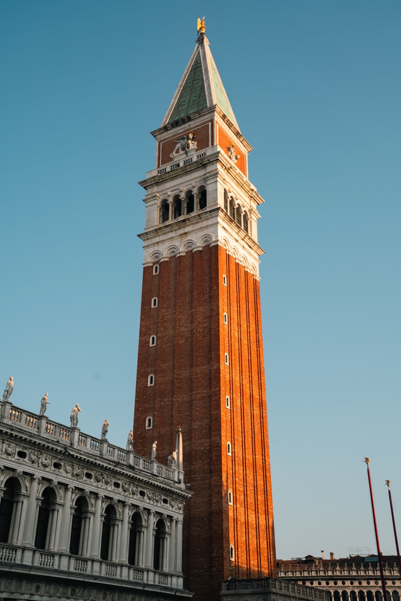 The Campanile of St Mark's rising against a blue sky, its brick shaft topped by a white belfry, gold lion and green pyramid roof, statues of the Marciana library at its foot