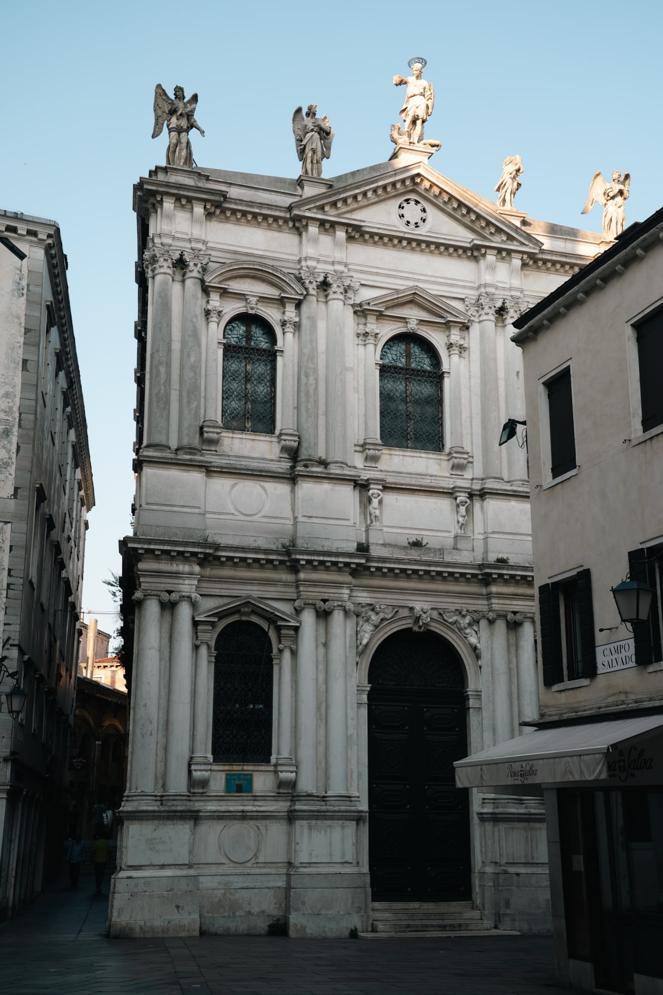 A white marble Baroque church facade in Campo San Salvador, winged angel statues standing along its roofline in the evening light