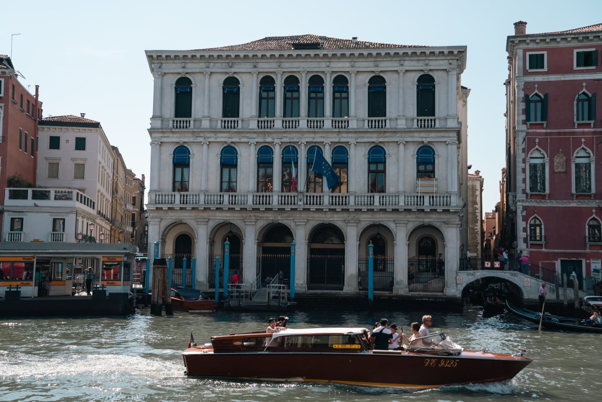 A varnished wooden water taxi passing a white arcaded Renaissance palazzo on the Grand Canal, a Rialto vaporetto pontoon at left