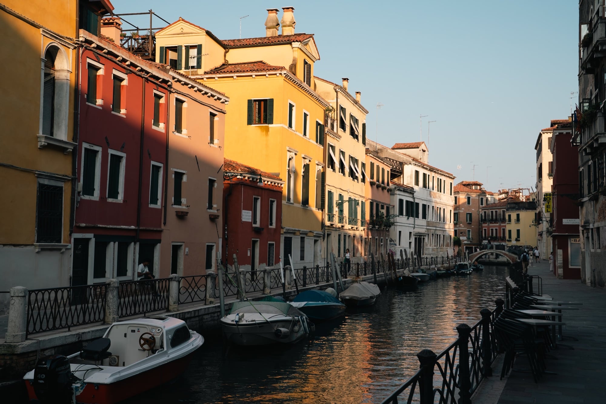 A quiet side canal lined with red, ochre and yellow houses, motorboats moored along the fondamenta beneath a small bridge