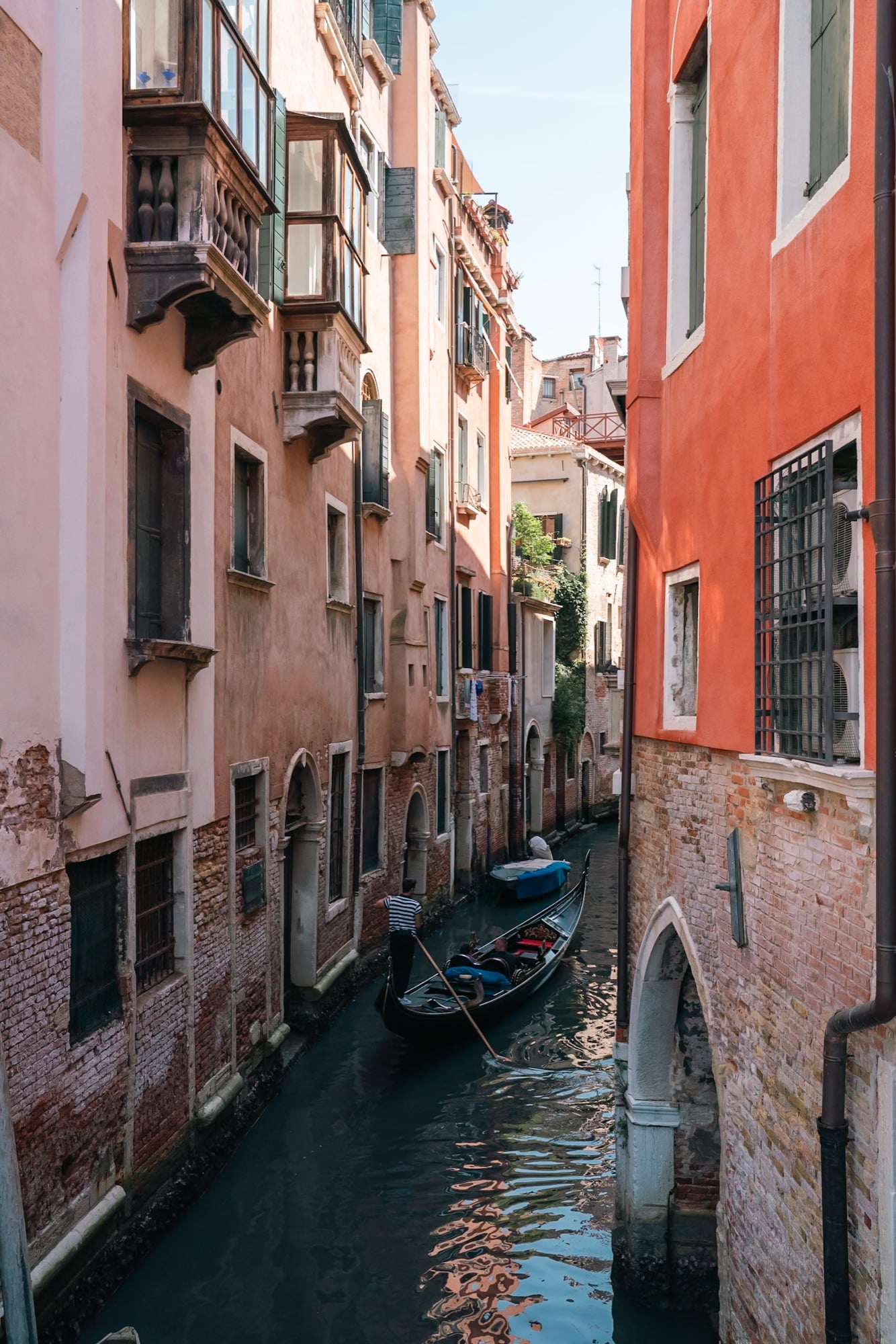 A gondolier poling an empty gondola down a narrow canal between weathered ochre houses with overhanging enclosed balconies