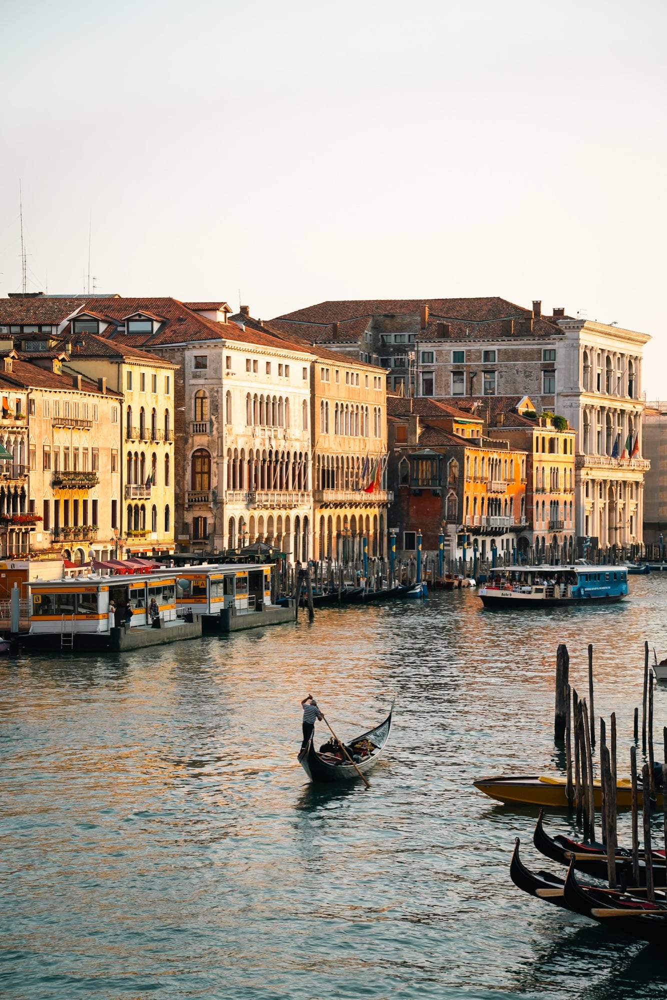 A lone gondolier rowing across the Grand Canal at golden hour, sunlit palazzi and the Rialto vaporetto stops lining the far bank