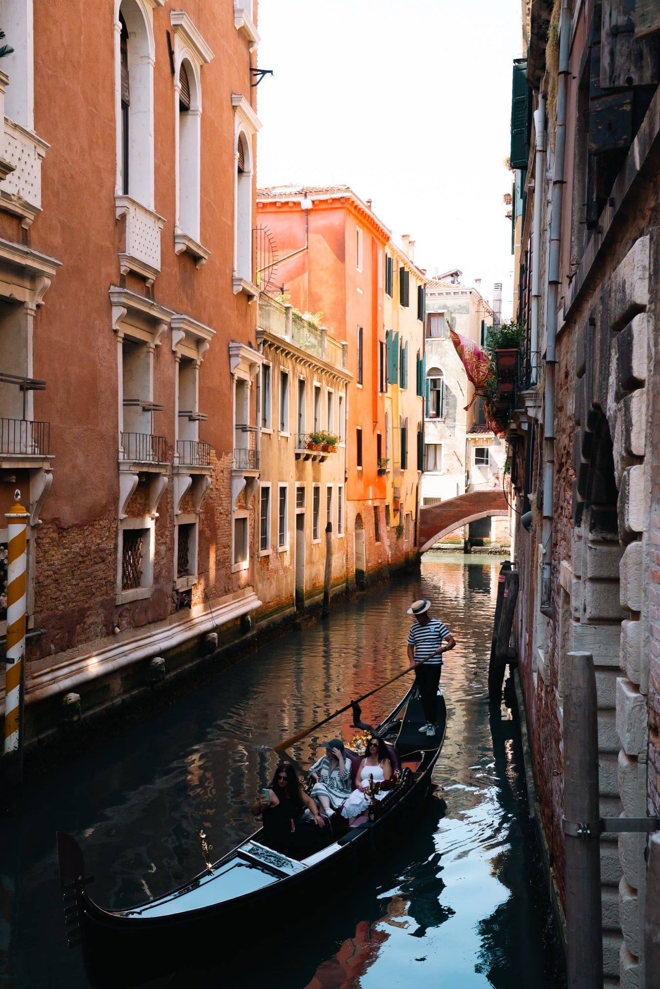 A straw-hatted gondolier rowing a gondola of passengers along a narrow canal between orange and terracotta buildings